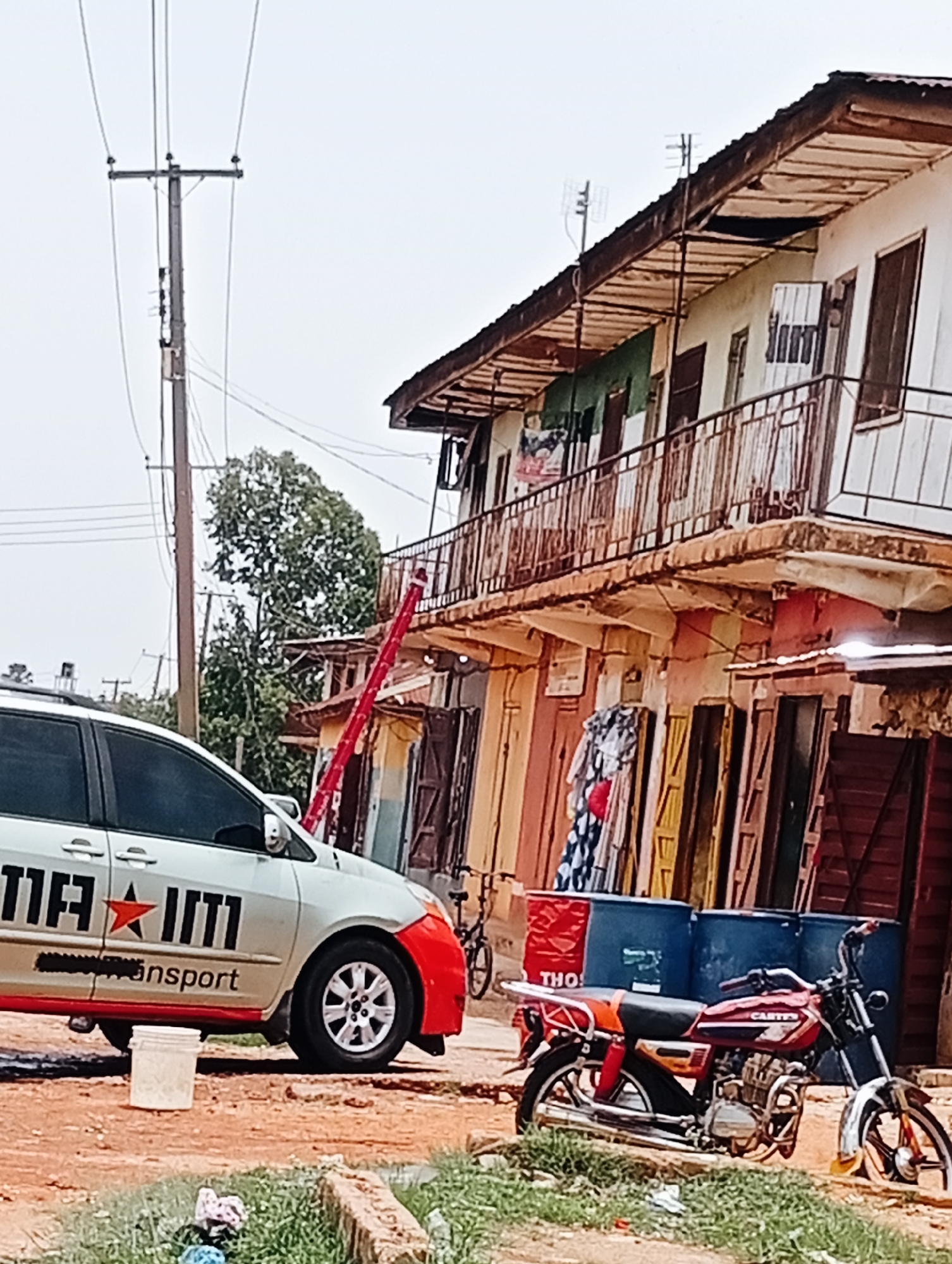 The image shows a street scene in Bukuru, Nigeria. A light-colored minivan with red accents and writing on the side is parked on a reddish-brown dirt road. A white bucket is next to the vehicle. A red motorcycle is parked behind the minivan, with three blue barrels visible behind it. In the background, a weathered two-story building with a balcony runs along the street. A red ladder is leaning against the building. Power lines are visible above the building. The sky is overcast.