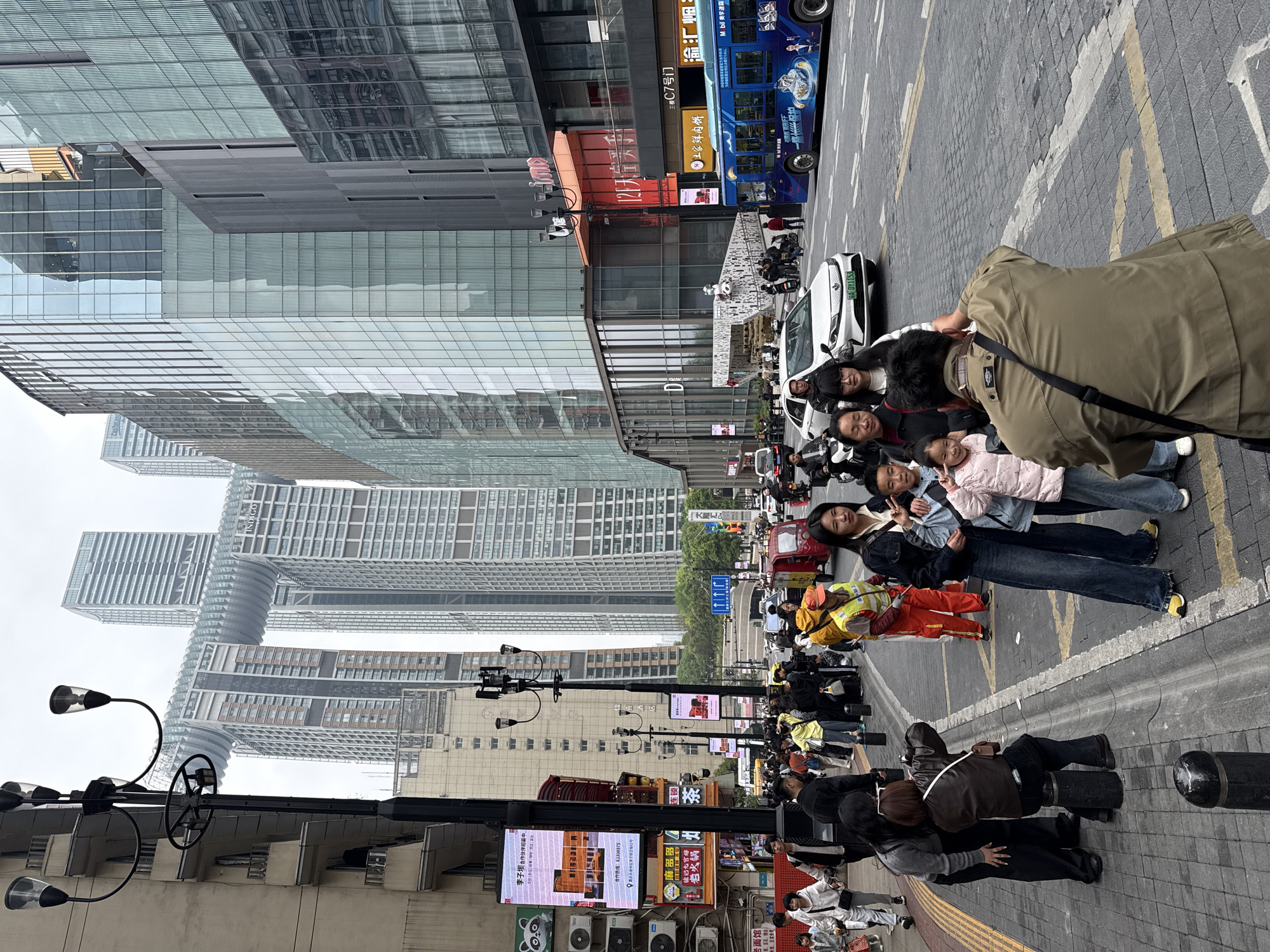 The image shows a bustling urban scene, likely in the Yuzhong District of Chongqing, China, judging by architectural and street signage characteristics. A group of people, possibly a family, are posing for a photograph on a pedestrian street. The photographer is visible in the foreground, capturing the group with his device. Behind them, a street with cars, taxis, and a bus can be seen. The backdrop consists of modern skyscrapers and other buildings, with storefronts and advertising displays along the lower levels. Street furniture such as lampposts and planters is also visible. Many other people are present, walking along the street.