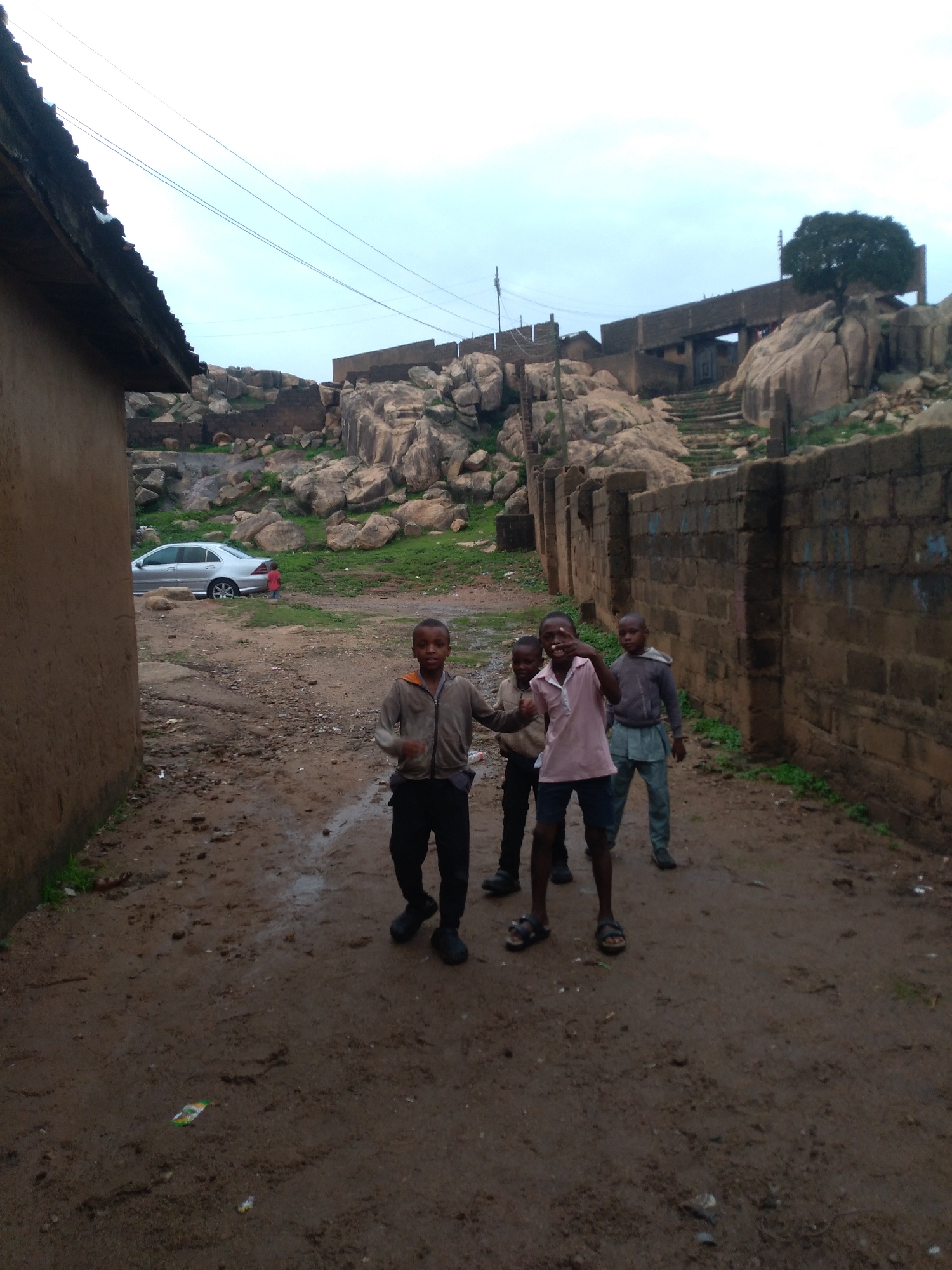 In the city of Jos, Nigeria, four young boys are pictured standing in a dirt pathway. The boy on the far left wears a gray zip-up jacket over black pants. The boy next to him is in a pink polo shirt and dark shorts. The third boy from the left is in a gray hooded sweatshirt and light blue shorts. The boy on the far right wears a gray hooded sweatshirt and blue pants. Behind the boys, a silver sedan is parked on the left side of the frame. The pathway is lined with a tall, rough-hewn brick wall on the right and a textured, mud-colored building wall on the left. Beyond the wall, a rocky hillside with scattered vegetation rises, leading up to structures built into the rock face. The sky is overcast, with a pale blue and gray hue.