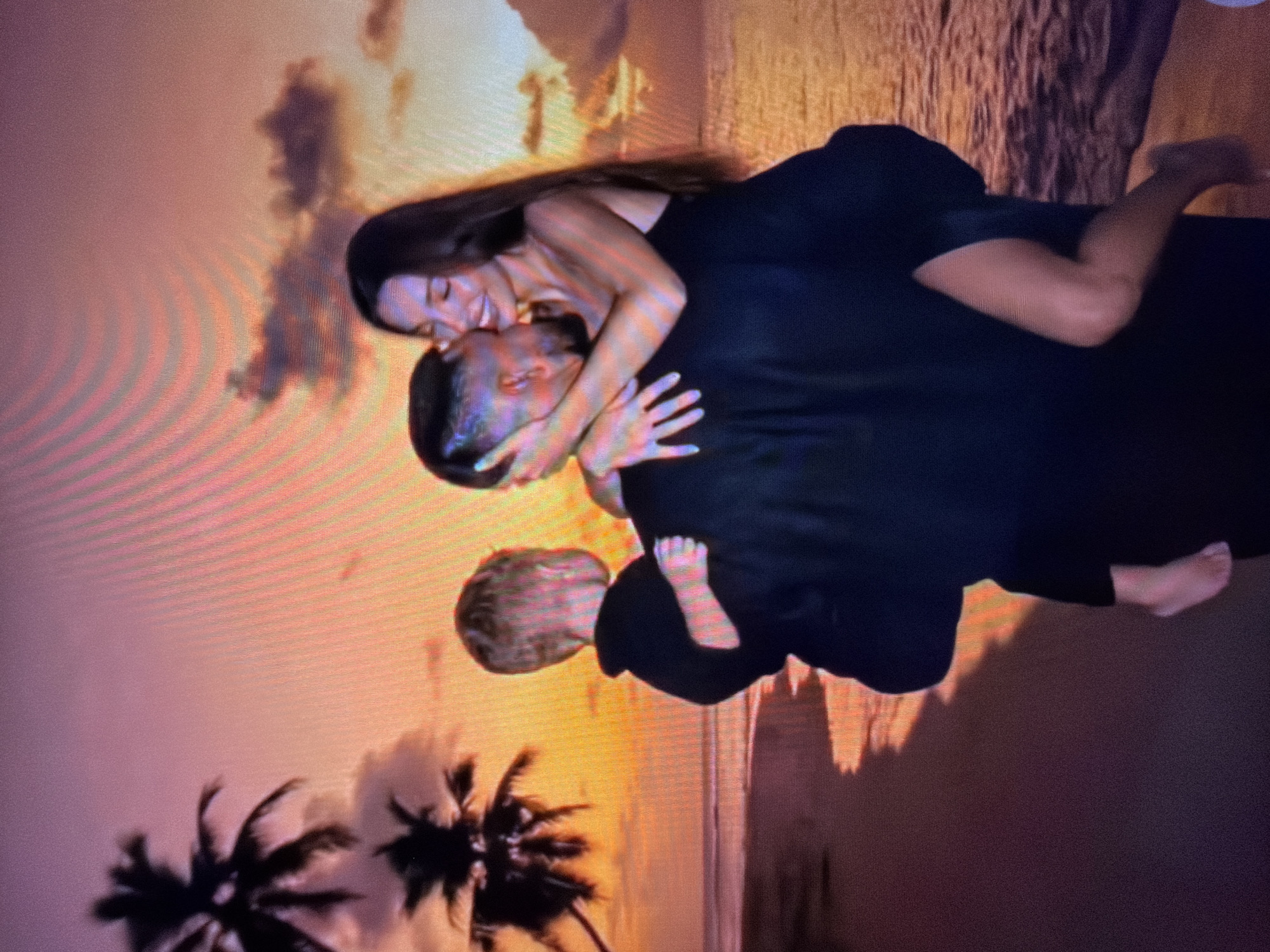 The image depicts a couple embracing on a beach at sunset, with a child held by one of the adults. The couple is dressed in dark clothing, and the woman is wearing a black dress. The background features palm trees, a body of water, and a vibrant sunset sky with clouds. The location is described as the city of Beșalma, Moldova.