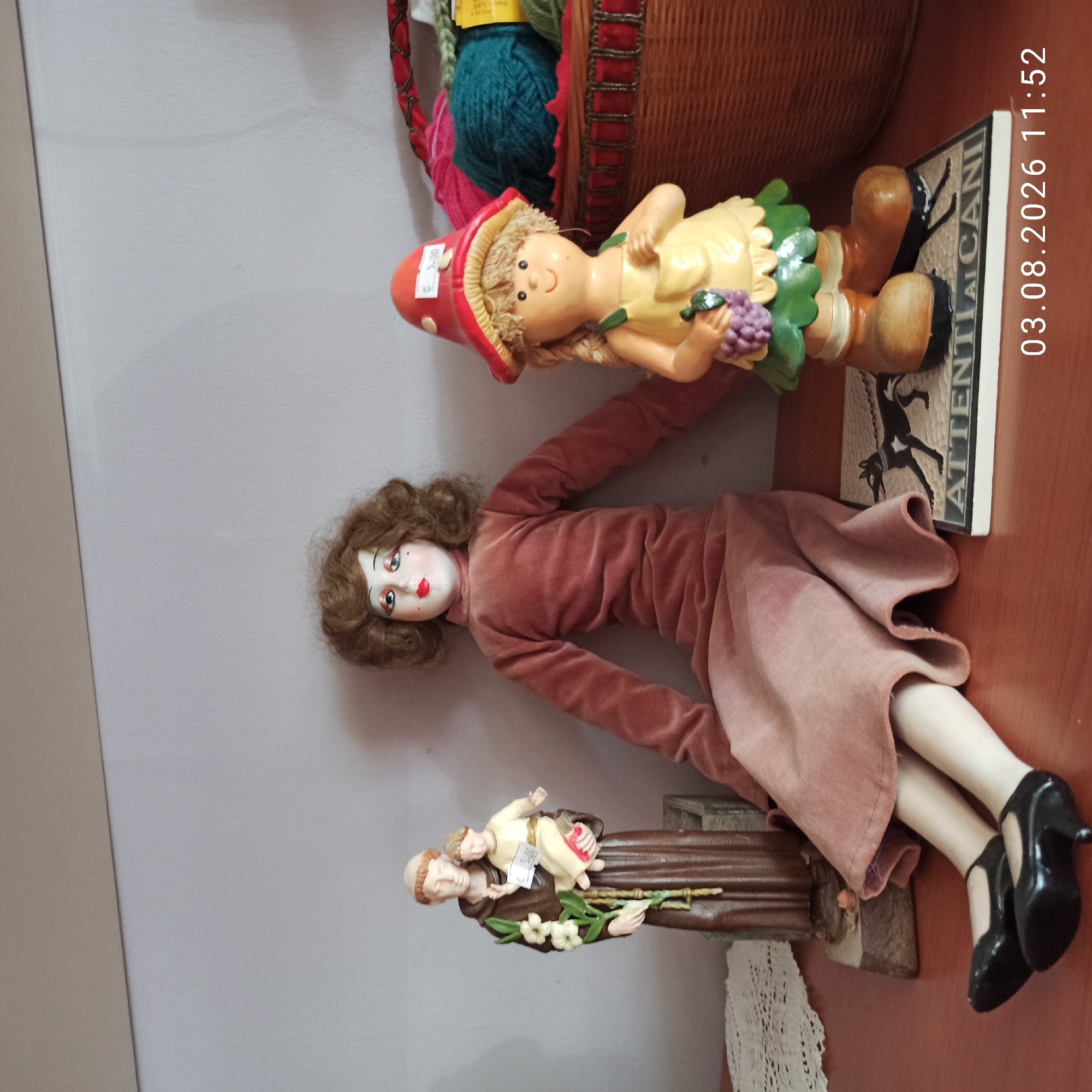 This media file displays a collection of decorative items, likely displayed in a shop or home in Ruvo di Puglia, Italy. The main subjects are two dolls and a religious figurine.
The first doll is a whimsical figure with a mushroom-shaped hat, yellow dress, and blonde braided hair, holding a bunch of grapes. It has a cheerful expression. Next to it, a larger doll is depicted lying down. This doll has detailed facial features with red lips and blue eyes, and is wearing a pink velvet dress. Its arms are extended, and its legs, clad in black high-heeled shoes, are crossed.
Below these dolls is a religious figurine of a saint, possibly Saint Anthony of Padua, holding a child. The figurine is brown and adorned with white flowers. A price tag is visible on the child's clothing.
In the background, a woven basket filled with colorful yarn and other items can be seen, along with a book or plaque that has text. The visible text on the plaque reads "ATTENTI AI CANI" which translates to "Beware of dogs". A timestamp "03.08.2026 11:52" is overlaid on the image, indicating the date and time the photo was taken. The lighting suggests it is daytime. The overall scene conveys a sense of curated display and perhaps a retail environment.