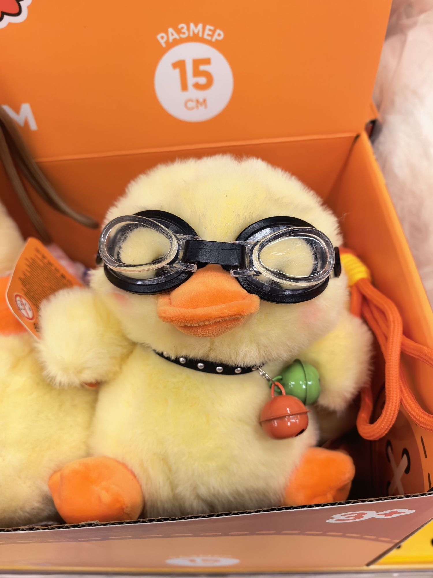 A plush yellow duck toy with large, clear goggles and an orange beak is sitting in an open orange box. The duck is wearing a black collar with silver studs and has two small bells attached to it: one green and one orange. The box has text indicating a size of "15 CM" and the word "РАЗМЕР" (Russian for "SIZE"). The background is blurred and appears to be a retail environment, possibly a toy store. The overall lighting is bright and suggests an indoor setting. There is no identifiable information or context linking this image to the city of Sterlitamak, Russia.