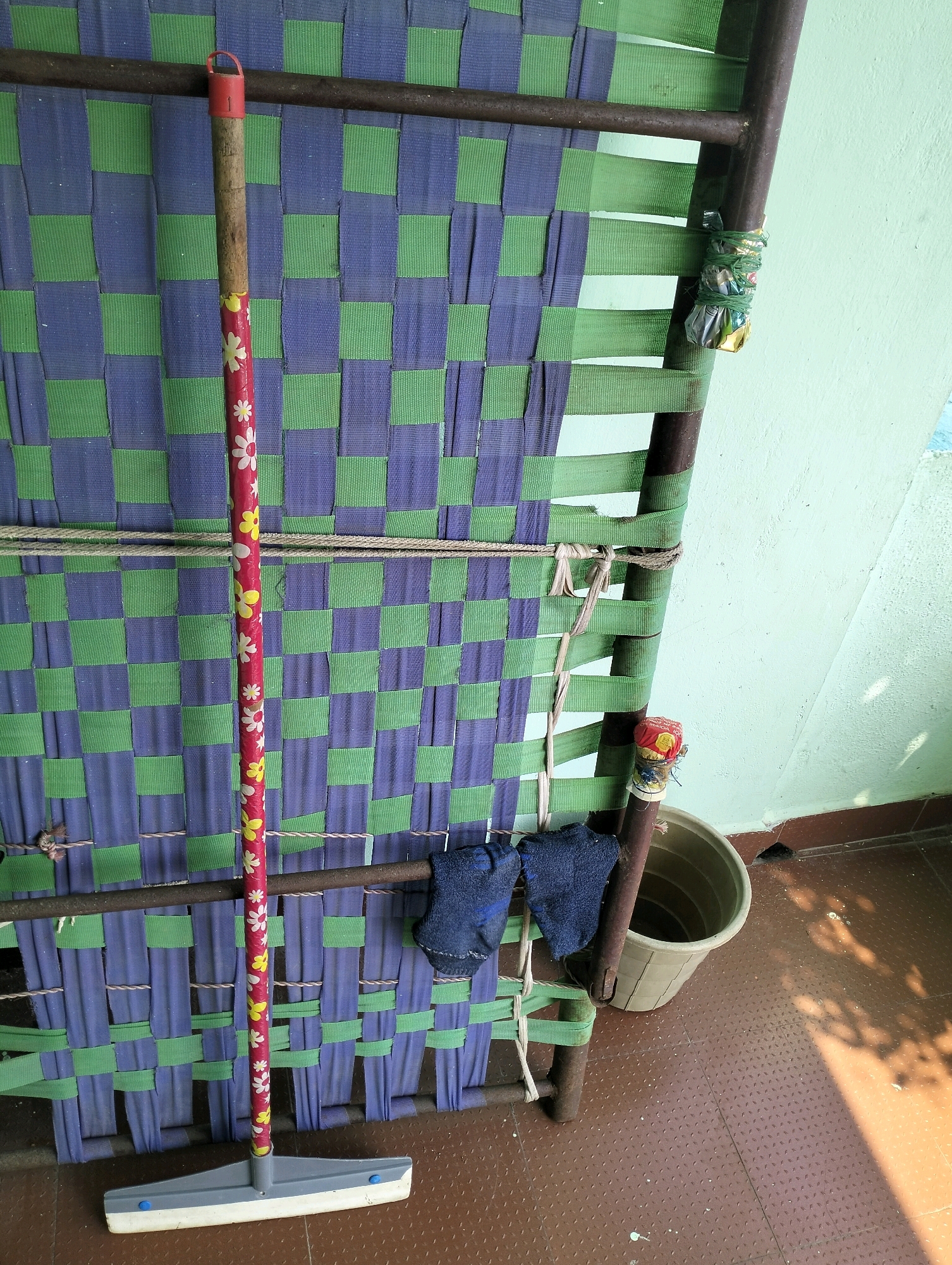 A floor squeegee with a red handle decorated with white and yellow flowers stands vertically, leaning against a cot with a woven mattress of alternating green and purple straps. Two dark blue work gloves are placed on the cot's frame, just above the mattress. To the right of the cot, a light brown plastic bucket sits on the floor. The floor is tiled with a brown, diamond-patterned material. The wall in the background is painted a pale green. The scene appears to be outdoors or in an open-air covered area, with sunlight casting shadows on the floor. The setting is consistent with a residential or storage area in Sriperumbudur, India.