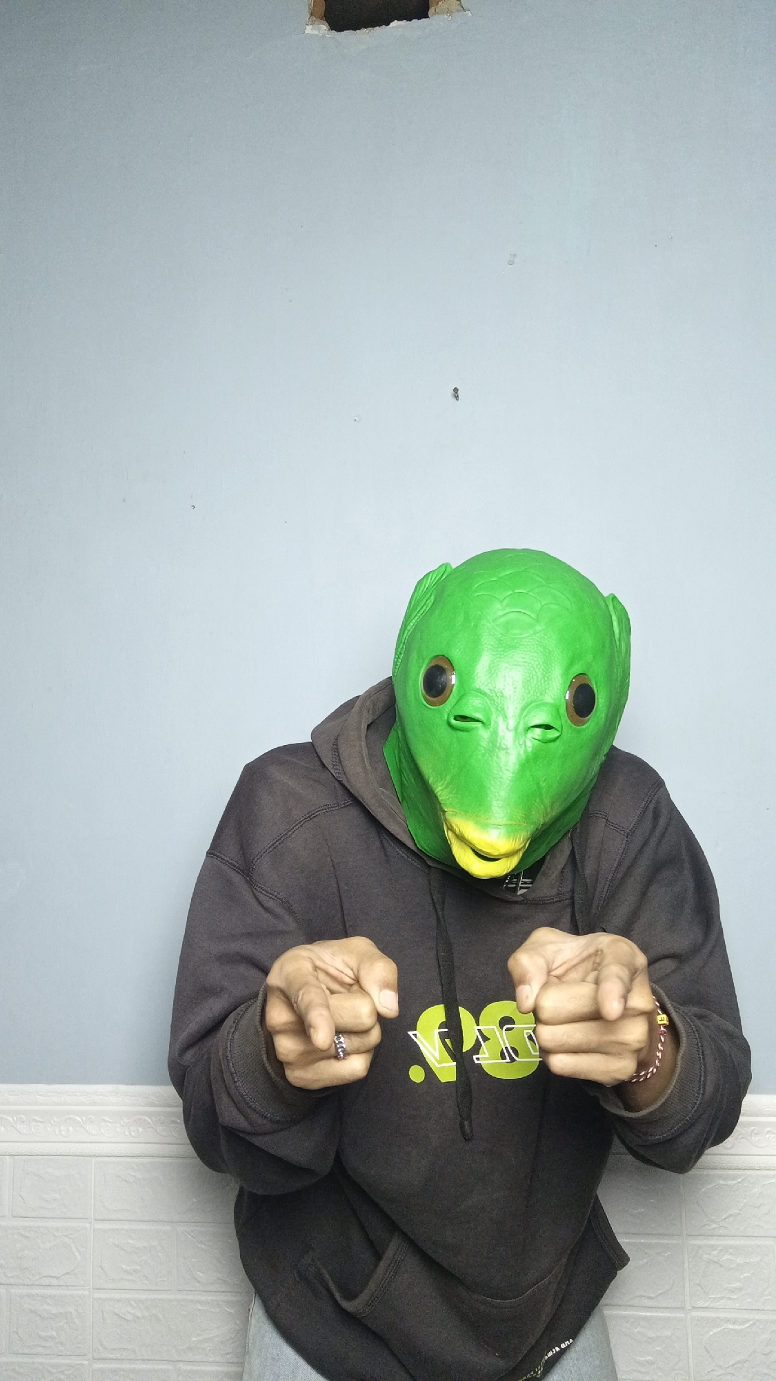 A person wearing a green alien mask and a dark gray hoodie stands in front of a pale blue wall. The mask has large, round, brown eyes and a yellow mouth. The person is extending their hands towards the camera, fingers pointed. The wall behind them has some damage near the top, and a white textured surface resembling brickwork runs along the bottom. This image was taken in Kebumen, Indonesia.