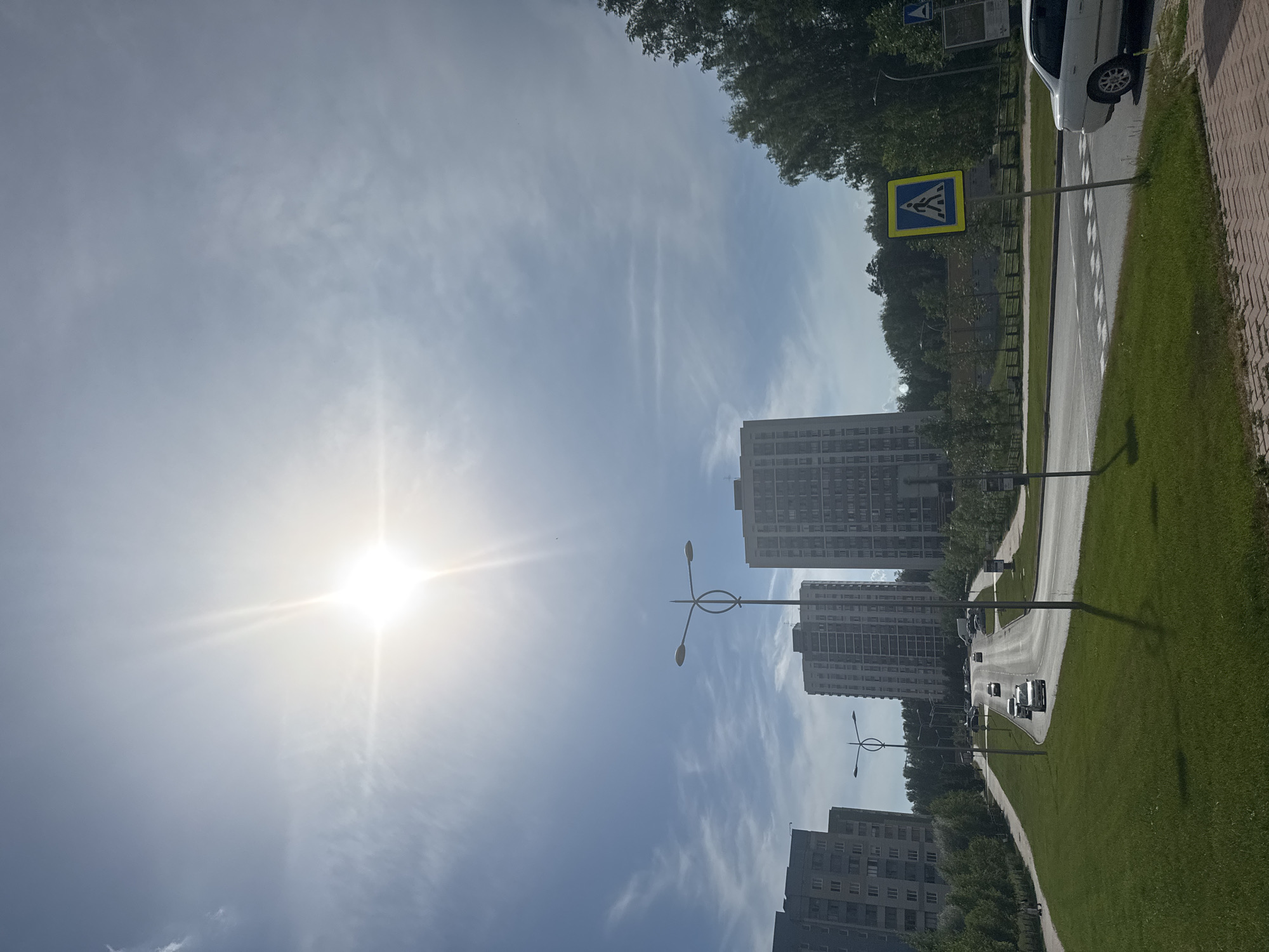 A sun-drenched scene captures a modern cityscape in Koltsovo, Russia, under a bright blue sky dotted with wispy clouds. The sun, a brilliant starburst, dominates the upper portion of the frame, casting a strong glare. In the foreground, a neatly mown grassy hill slopes down towards a paved road. The road curves gently upwards, with several cars driving on it.
Tall, multi-story apartment buildings line the sides of the road, their modern architecture standing out against the natural backdrop of green trees. Streetlights with distinctive oval designs are strategically placed along the road, illuminating the urban environment. A yellow pedestrian crossing sign with a silhouette of a person walking is prominently visible on the right side of the road, indicating a crosswalk. A white car is parked near the pedestrian crossing. The presence of these elements suggests a well-developed residential area with functional urban infrastructure. The overall impression is one of a peaceful, everyday scene in a contemporary Russian city.