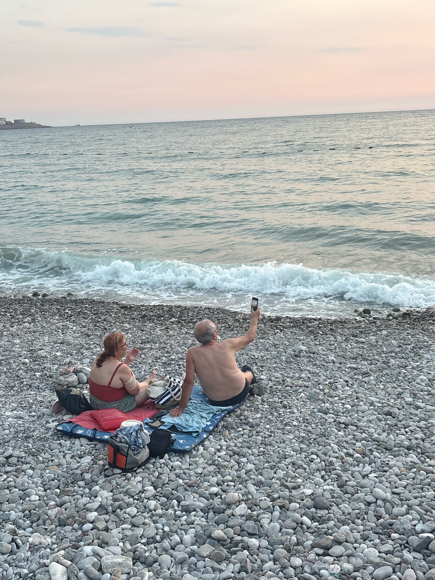 An elderly couple is having a picnic on a pebble beach in Bar, Montenegro, near the Adriatic Sea. The man is wearing dark shorts and is shirtless, holding up a smartphone to take a selfie. The woman is wearing a red swimsuit and patterned shorts, and is sitting next to him. They are sitting on blankets spread out on the pebble beach. Bags and a cooler are present near the couple. The sea is calm with gentle waves rolling onto the shore. The sky is overcast with a light pink hue, suggesting it is either sunrise or sunset. In the distance, there are some buildings on the left side of the frame, and a faint line of buoys in the water.