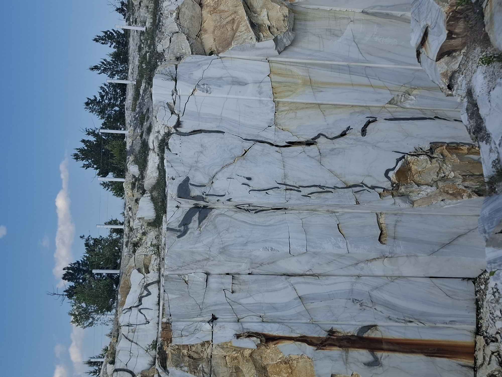 The image shows a quarry with large blocks of light-colored stone, likely marble, with dark veining. The quarry is situated in a rocky, uneven terrain with some vegetation and trees visible in the background. A white fence runs along the edge of the quarry. The sky is blue with some white clouds. The stone blocks have a rough, fractured surface, indicating they have been recently quarried. The overall impression is one of a natural landscape altered by industrial activity.