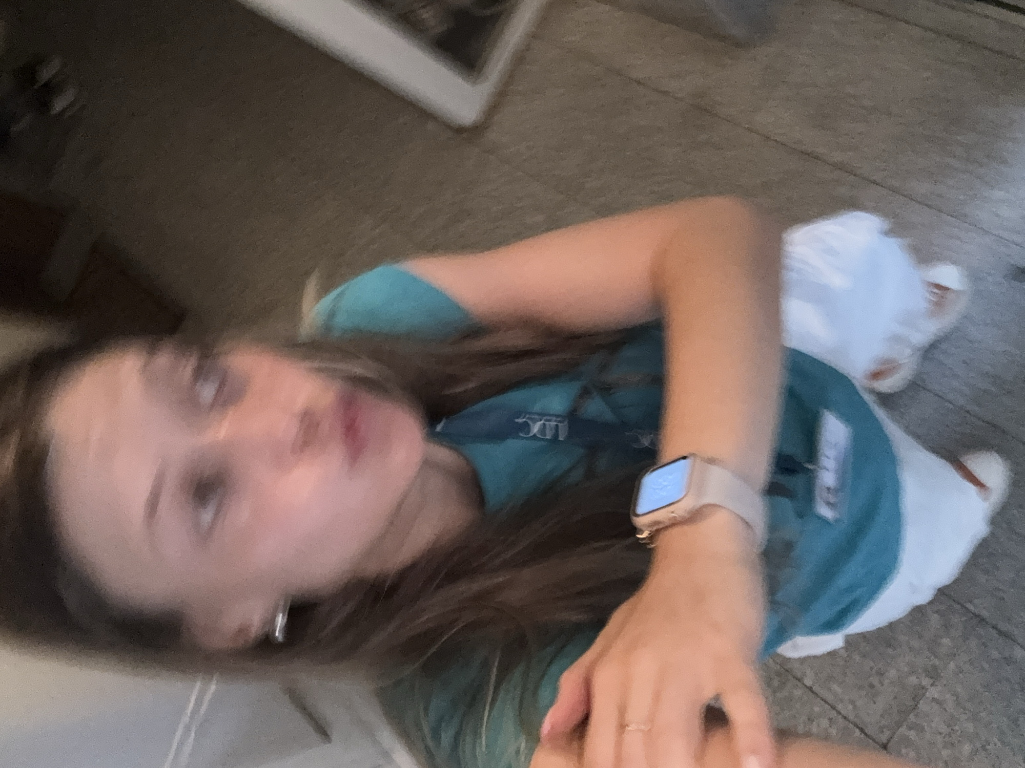The image is a blurry, low-angle selfie taken indoors, likely in a residential setting. A young woman with long brown hair is in the foreground, looking towards the camera with her lips pursed. She is wearing a teal t-shirt with a lanyard that has a white badge clipped to it, and white shorts. On her left wrist, she wears a light-colored smartwatch. Her right arm is bent at the elbow, with her hand resting near her chest. The background is out of focus, showing a floor with what appears to be tile or wood planking and indistinct furniture or wall elements. There are no visible clues to indicate the specific location within the city of Sofia, Bulgaria, or any urban context. The overall impression is of a casual, candid moment.