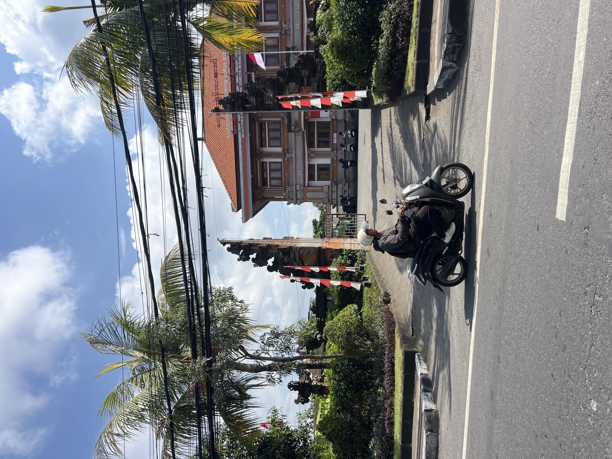 A person wearing a helmet and dark clothing rides a dark-colored scooter on a paved road in Denpasar, Indonesia. The scooter is in the foreground, slightly to the left of the frame, and facing towards the right. Behind the scooter, a traditional Balinese gate with intricate stone carvings stands to the left, adorned with red and white striped flags. Beyond the gate is a multi-story building with an orange tiled roof, a common architectural style in the region. Lush green vegetation, including palm trees and manicured bushes, surrounds the gate and the building. The sky above is blue with scattered white clouds. The image captures a typical street scene in Denpasar, reflecting the island's cultural heritage and tropical climate.