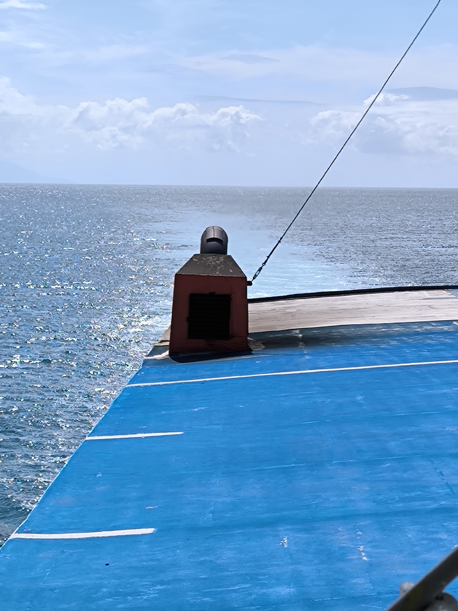 The image shows a view from the deck of a ship, moving across the ocean. The deck is painted bright blue with white stripes. A dark red structure with a black top, possibly a vent, stands near the edge of the deck. A cable runs diagonally from the top right of the image towards the structure. The ocean stretches out to the horizon under a sky with scattered clouds. A faint outline of land is visible in the distance. The location is Lembar Selatan, Indonesia.