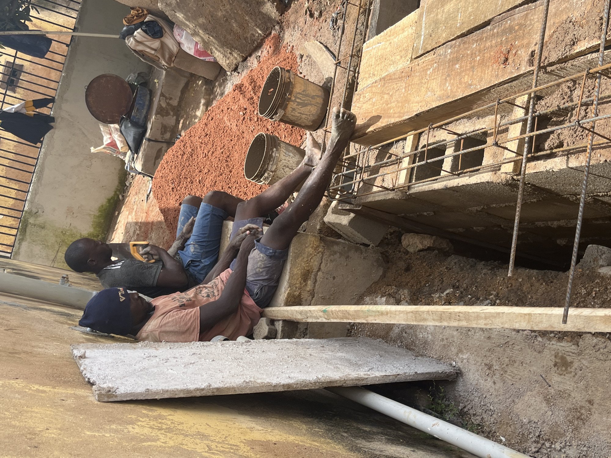 Two individuals are present at a construction site. The individual on the left is wearing a dark blue beanie, an orange t-shirt, and dark blue shorts. They are sitting with their legs extended, resting against a rough, light-colored rectangular object leaning against a yellow-colored wall. The individual on the right is wearing a dark t-shirt with a pattern and blue denim shorts. They are also sitting, with their hands in their lap and holding a yellow object.
The environment appears to be an outdoor construction area. In the background, there are piles of reddish-brown material, possibly soil or gravel, and several buckets. There are also some bags, a large round metal object, and items of clothing hanging on a metal fence. In the foreground, there is an excavated trench with rebar and wooden planks. A white pipe is visible on the left side of the image. The overall scene suggests a construction project in progress. The location is identified as Jos, Nigeria.
