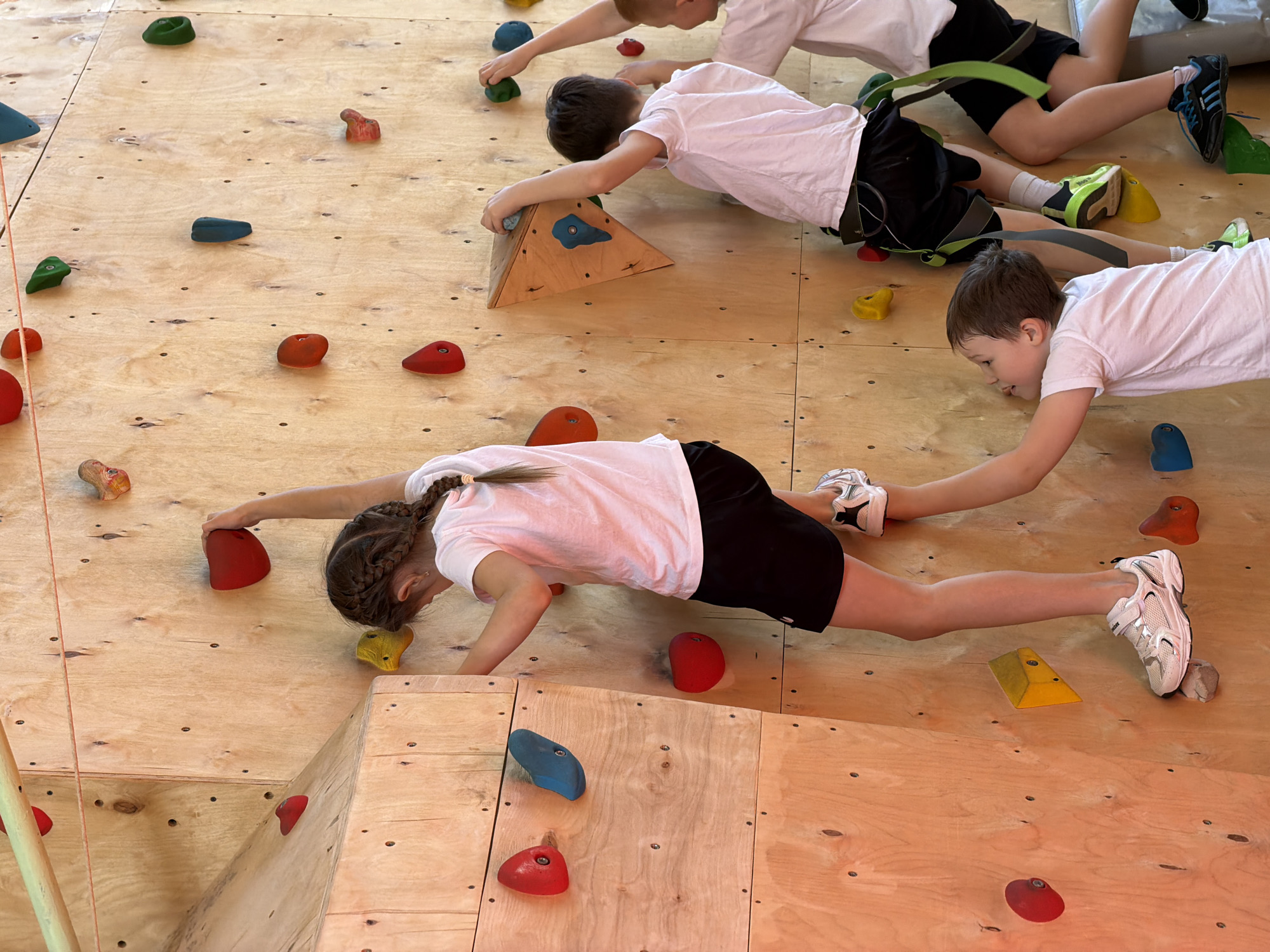 A high-angle shot depicts four children on an indoor climbing wall, likely in a recreational facility. All four children are wearing white t-shirts and dark shorts, positioned horizontally against the light-colored wooden wall. The wall is fitted with colorful climbing holds of varying shapes and sizes. One child, a girl with braided hair, is in the foreground, extending her arms and legs to grip the holds. Two other boys are positioned above her, and a fourth child is partially visible at the top. One of the boys wears a harness. The location is presumed to be in Izhevsk, Russia.