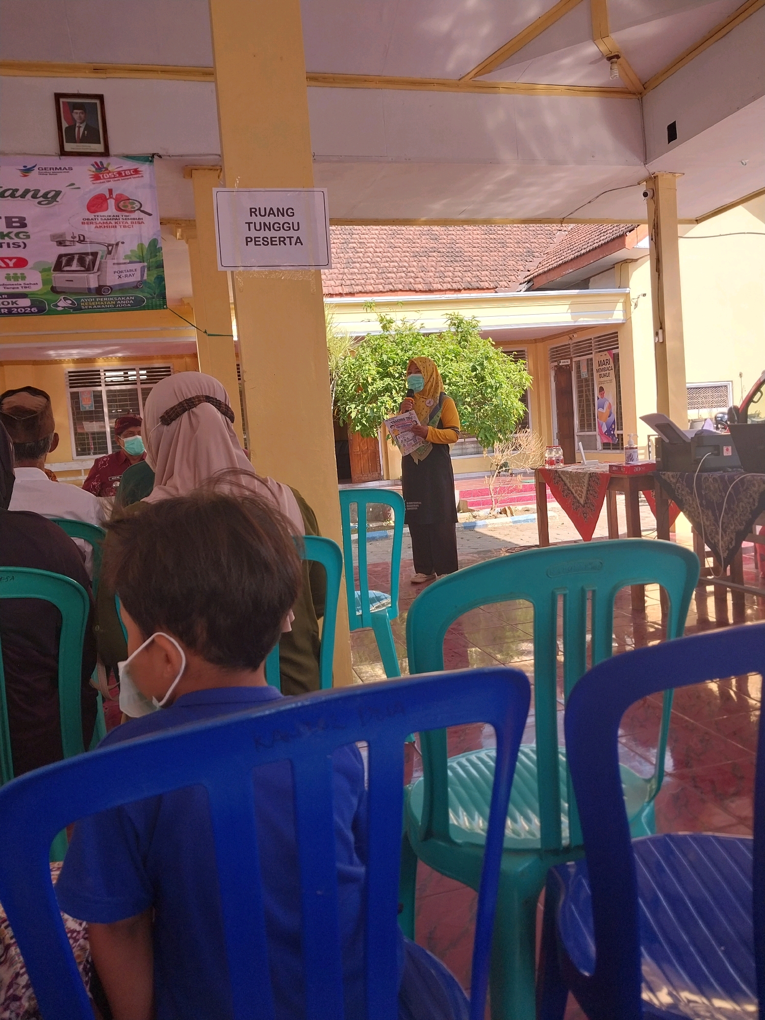 The image depicts an outdoor gathering, possibly a health seminar or community event, taking place in Bangsri, Indonesia. In the foreground, several blue and teal plastic chairs are visible, some occupied by individuals. A young boy wearing a blue shirt and a face mask is seated in a blue chair, facing away from the camera. Further back, a woman in a yellow hijab and black attire stands and addresses a group of people, holding a pamphlet. She is positioned near a table with various items, including a printer and hand sanitizer. Behind her, a banner with Indonesian text and an image of an X-ray machine is displayed. The background reveals a building with tiled roofs and trees, suggesting a public facility or community center. The overall atmosphere appears organized and participatory.