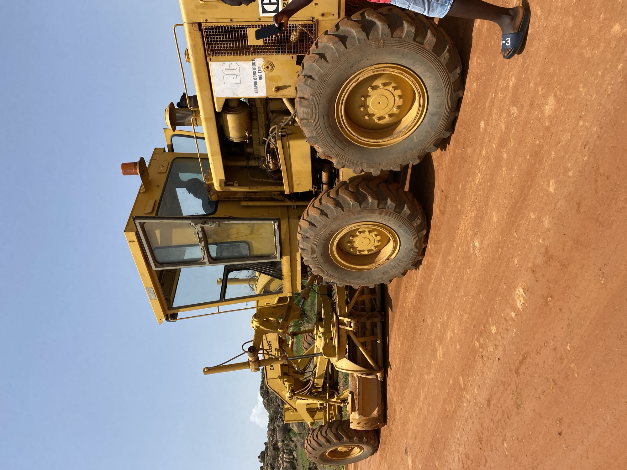 This media file shows a yellow grader, a type of heavy construction equipment, parked on a reddish-brown dirt surface under a clear blue sky. The grader is positioned at an angle, with its large tires and heavy machinery clearly visible. The scene appears to be an outdoor construction site or an area where such equipment is stored.
A person, partially visible, is standing near the grader. They are wearing blue shorts and a red shirt, with dark skin. One of their feet is visible, wearing a black sandal with "1-3" written on it. The person is holding a black smartphone, suggesting they might be taking a picture or using it for some other purpose.
Notable details include the "EC" logo and the text "ERAPON CONSTRUCTION N.I.G. LTD" on a white label attached to the grader's body. The lighting suggests it is daytime, likely a sunny day given the clear sky and the shadows cast. In the background, there is a hint of rocky hills, possibly indicating a semi-arid or hilly terrain. The overall impression is a functional, operational environment associated with construction or roadwork in Bukuru, Nigeria.