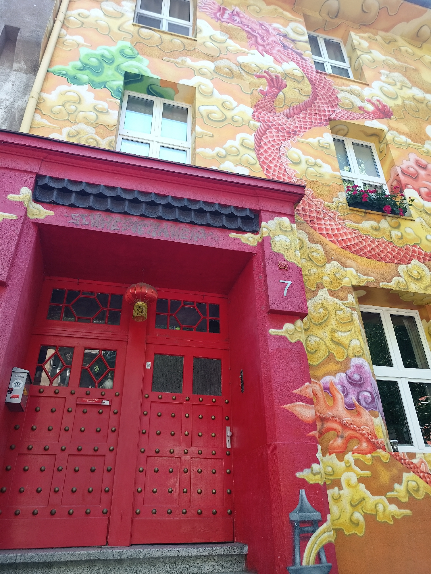 The image shows the facade of a building in Düsseldorf, Germany. The building features a vibrant mural of a pink dragon surrounded by yellow and orange clouds, painted in a traditional Chinese style. A bright red entrance with a decorative lantern hangs above the double doors. The doors themselves are red and adorned with numerous brass-colored studs. Above the entrance, a tiled roof-like structure is visible. White-framed windows are scattered across the building's facade, some with small window boxes filled with pink flowers. A red mailbox is mounted on the left side of the entrance. The building is situated on a street, with a gray stone step leading up to the entrance. The overall impression is of a building with a distinctly Asian-inspired aesthetic.