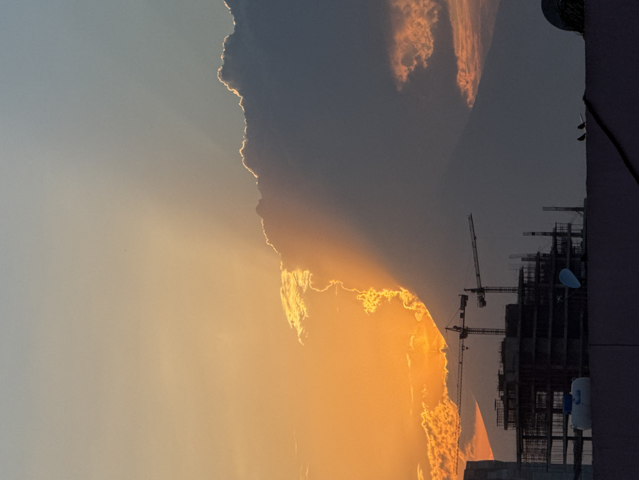 The image displays an urban scene at dusk or dawn in the city of Puebla, Mexico. The sky occupies the upper two-thirds, exhibiting a pronounced contrast. The left side is illuminated by an intense orange-yellow glow, indicating direct sunlight, while the right side is a darker blue-grey. A large, dark cloud formation dominates the right half of the sky; its left edge is brightly outlined in vivid orange and yellow where sunlight strikes it. Faint crepuscular rays emanate from behind this cloud. Further to the right and lower, smaller, reddish-orange clouds are visible against the darker sky.
In the mid-ground, the silhouette of a large, conical mountain, consistent with the Popocatépetl volcano, is discernible, rising behind an urban construction zone. The construction site features a multi-story building under development, composed of a dark, skeletal frame with visible rebar and structural elements. Two tall tower cranes are silhouetted against the bright sky, positioned above and behind the building frame.
The immediate foreground is a dark, flat rooftop or parapet wall. On this surface, from left to right, are a white, cylindrical water tank, a blue satellite dish, and two small, dark birds perched on a thin, horizontal element, possibly a wire or pipe. A dark, diagonal line, likely a cable or pipe, crosses the rooftop from the lower-left to the mid-right. No human figures are present.
