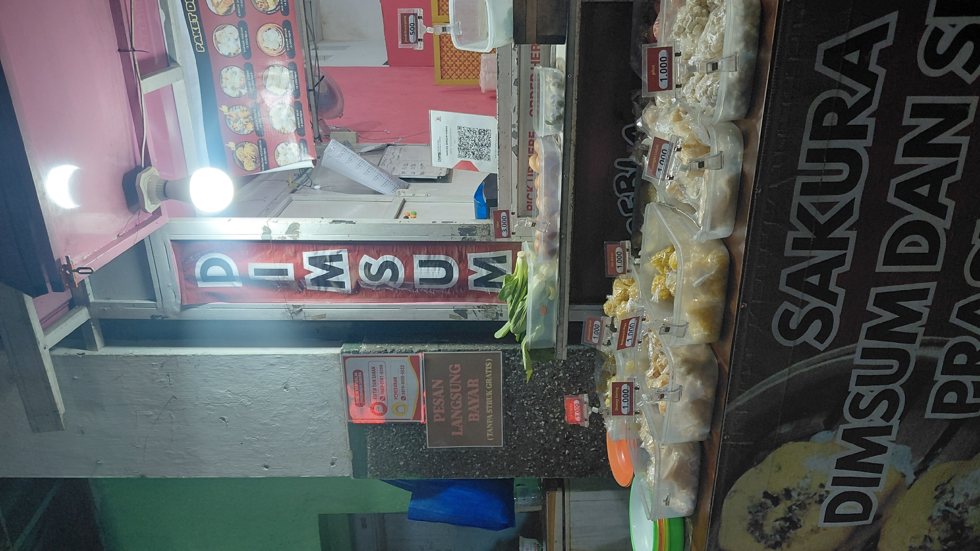 The image shows a street food stall in Bantarsoka, Indonesia, selling dim sum. The stall is brightly lit by an overhead lamp, casting shadows on the surrounding area. A large vertical banner behind the counter reads "DIMSUM" in red letters. Various containers of dim sum are displayed on the counter, with price tags of 1.000 and 3.000 Indonesian Rupiah visible. A sign on the left side of the stall instructs customers to "PESAN LANGSUNG BAYAR (TANPA STRUK GRATIS)", meaning "ORDER DIRECTLY PAY (FREE WITHOUT RECEIPT)". Another sign above it indicates "KRITIK DAN SARAN" (Criticism and Suggestions) and provides a phone number. A QR code for payment is also displayed. In the background, a menu with images of dim sum and prices is visible. The overall scene depicts a typical night market or street food vendor setup.