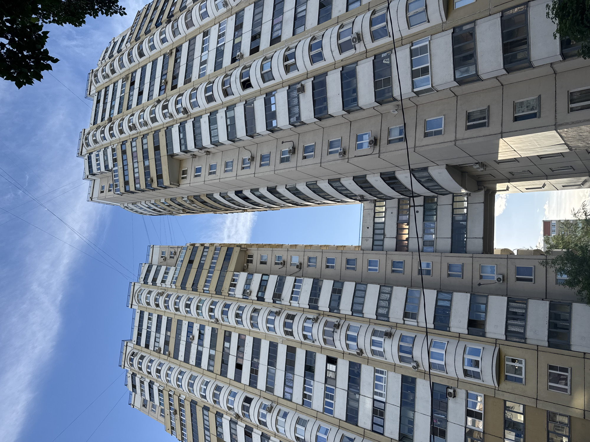 This is a low-angle shot of two residential apartment buildings in Moscow, Russia. The buildings are primarily concrete and white, with numerous windows and curved balconies. The sky is blue with wispy clouds visible in the space between the structures. There are some trees at the edges of the frame. Power lines are visible against the sky.