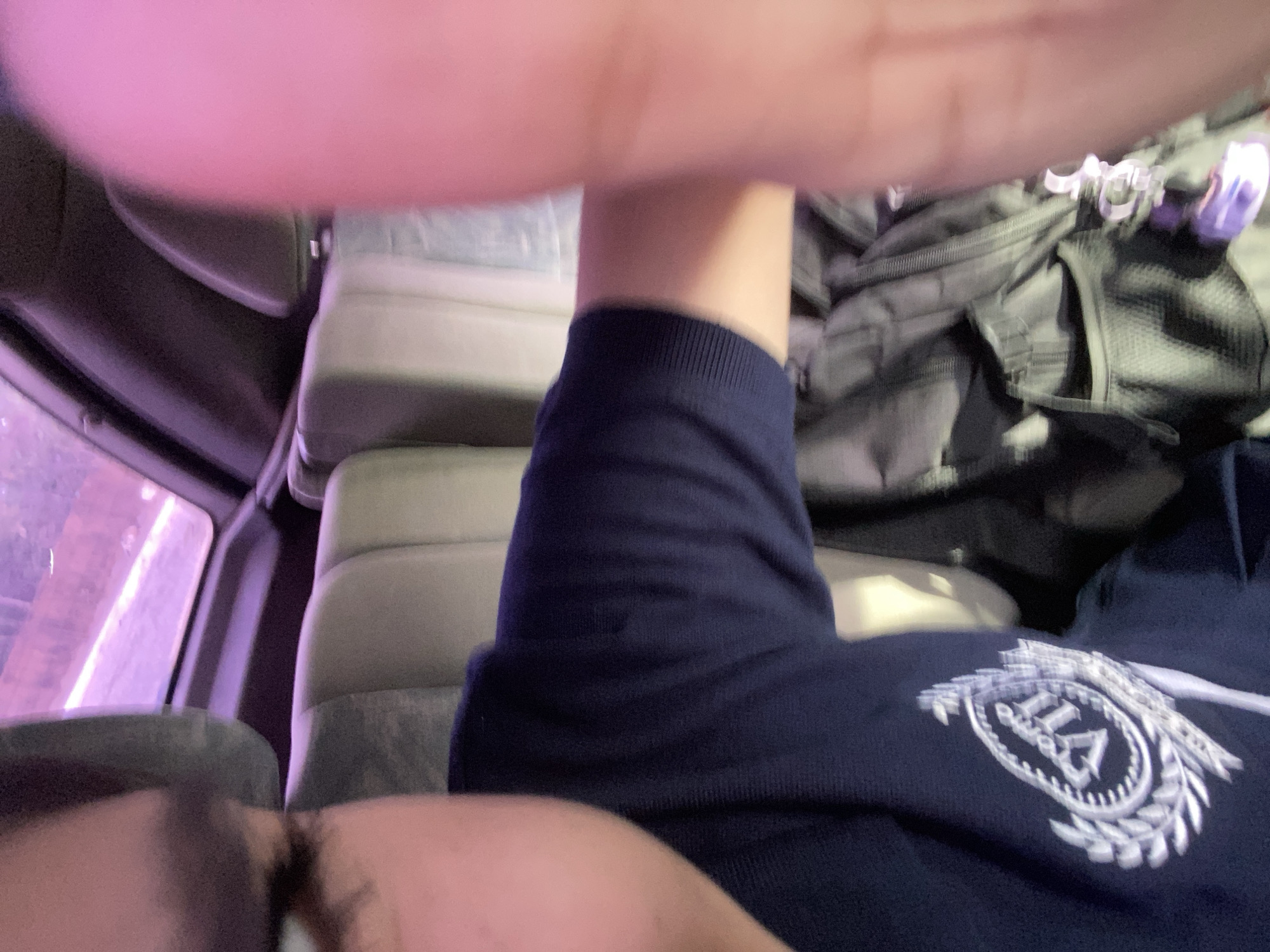 The image shows a person sitting in a vehicle, possibly a bus, with a partially visible face at the bottom. The person is wearing a navy blue garment with a white emblem. Behind the person are cushioned seats and a dark gray backpack. A window is visible to the left. There is no evidence in the picture to confirm a location within the city of Cotoperíz, Venezuela.