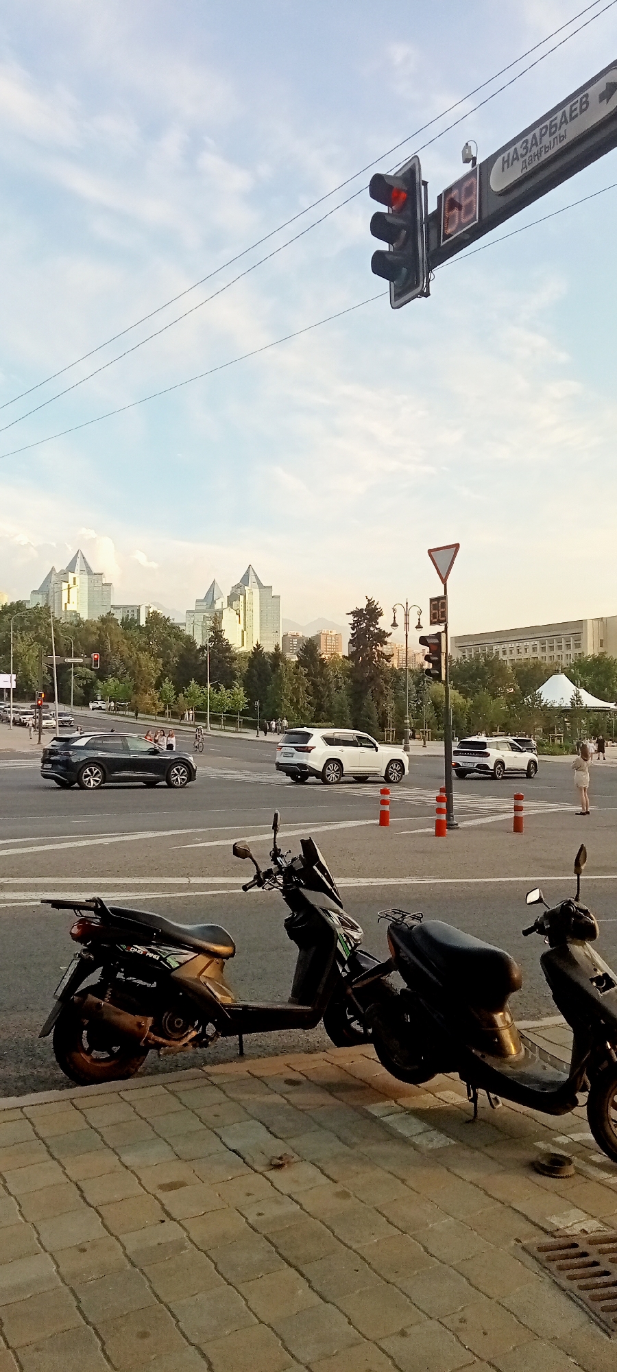This is a street-level, full-frame photograph taken in Almaty, Kazakhstan, likely on Nazarbayev Avenue, given the prominent street sign. The image captures a street intersection with traffic lights, road markings, and several vehicles. In the foreground, two black scooters are parked on a patterned sidewalk, facing the street. One scooter is mostly visible, while the other is partially obscured.
The middle ground shows a busy intersection with cars moving. A dark-colored SUV is on the left, turning, while two white SUVs are positioned further into the intersection, suggesting they are stopped or proceeding. Several orange traffic cones are visible on the road. Pedestrians can be seen on the right side of the intersection.
In the background, distinctive modern buildings with pointed roofs, characteristic of Almaty's architecture, rise above a dense line of green trees. The sky is partly cloudy, with a pale blue hue and wispy clouds, indicating it is likely late afternoon or early evening. A traffic light with a red signal is illuminated. A digital display showing the number "69" is attached to the traffic light pole. The overall scene depicts a typical urban environment in Almaty during daytime.
