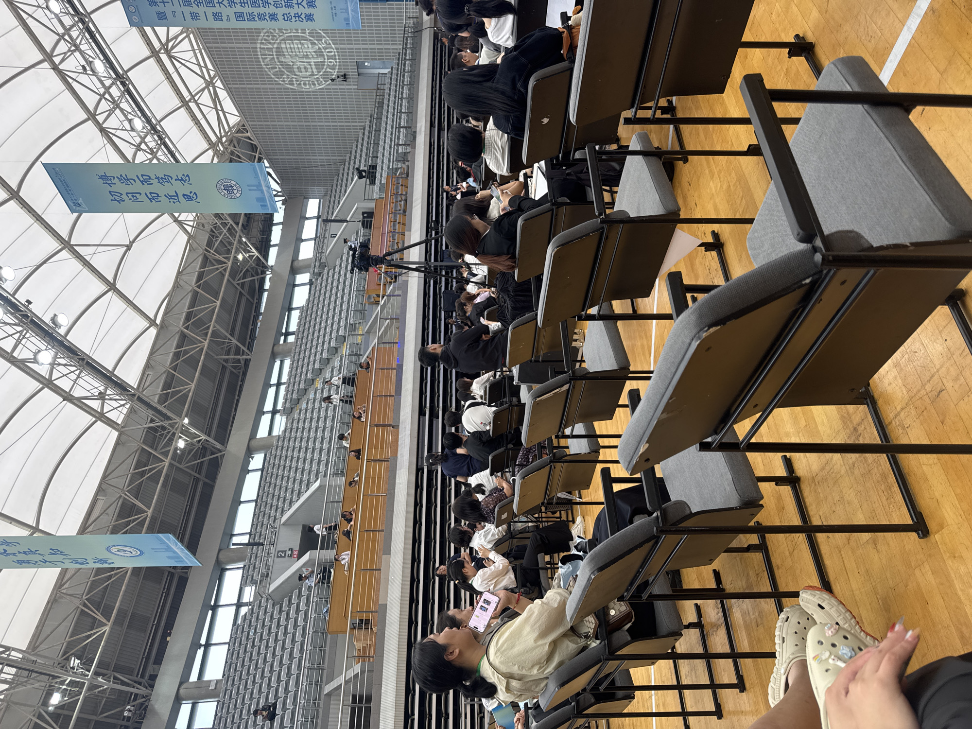 The image depicts an indoor event or lecture taking place in a large auditorium. Rows of grey stadium seating are visible in the background, mostly empty. In the foreground and middle ground, people are seated in chairs arranged on a wooden floor. Many attendees are looking towards a stage area, though it is not fully visible. Some individuals are using their mobile phones. A professional video camera on a tripod is positioned on the left side of the frame, suggesting a recording or broadcast is in progress. Several large banners with Chinese characters are hung from the ceiling. The environment appears to be a university campus or a public event space, possibly in the Yangpu District of China, given the presence of Chinese text and architectural style. The lighting is artificial, coming from overhead fixtures.
