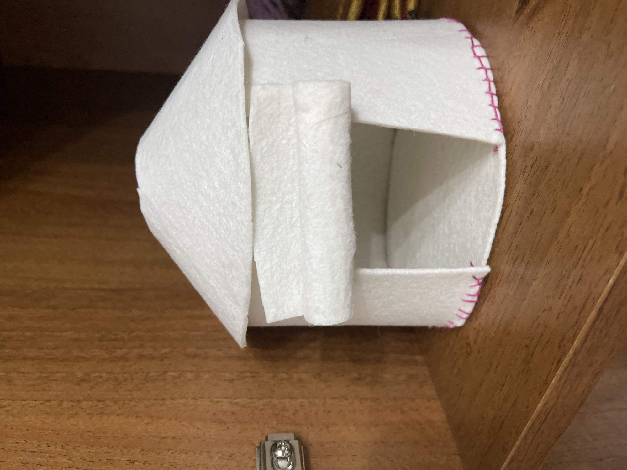 A white felt yurt-shaped dollhouse sits on a wooden shelf. The yurt has a conical roof and a semicircular opening at the front. A flap of felt hangs over the opening, rolled up on one side. The bottom edge of the yurt is stitched with pink thread. The background is a wooden cabinet with vertical grain. A metal hinge is visible on the left side of the frame. The scene is indoors.