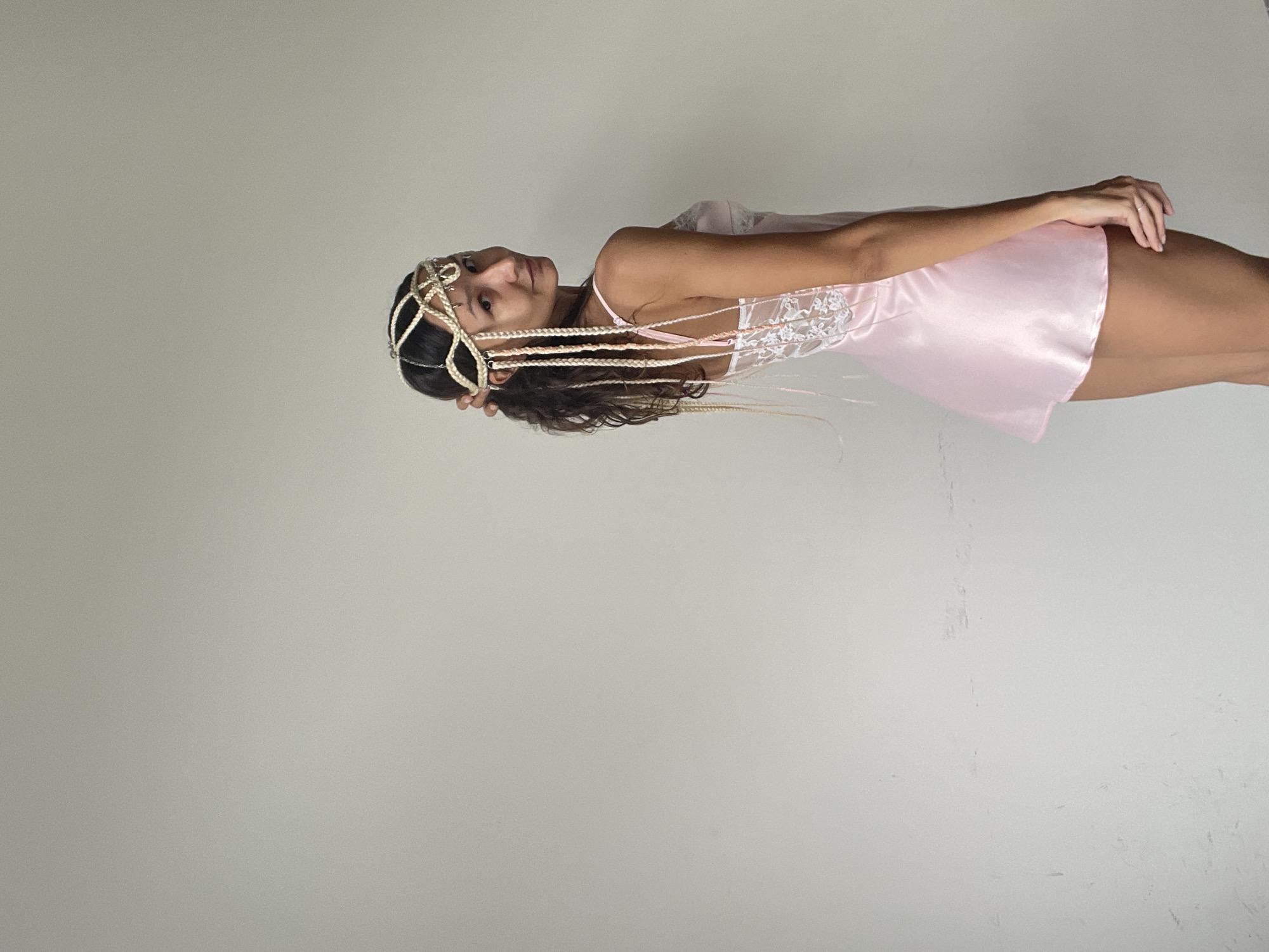 A young woman with dark, curly hair adorned with a decorative headpiece stands against a plain, light-colored wall. She is wearing a short, pale pink satin slip with white lace detailing on the bodice. Her hair is styled with braids that hang down, some interspersed with thin ribbons. She is looking over her shoulder at the camera with a slight smile. The lighting suggests an indoor setting. The image contains no elements that suggest a location in Mũi Né, Vietnam.