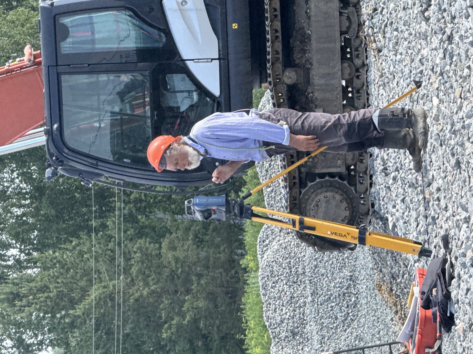 In the city of Leninsk, Russia, a man wearing an orange hard hat, blue collared shirt, and gray pants is operating a surveying instrument. The instrument is mounted on a yellow tripod, and the man is looking through its eyepiece. He has a long, white beard and is wearing black boots. He is standing on a pile of gravel, next to a large black excavator. The excavator's tracks are visible in the foreground. In the background, there are green trees. A red toolbox with some equipment is on the ground to the left of the man.