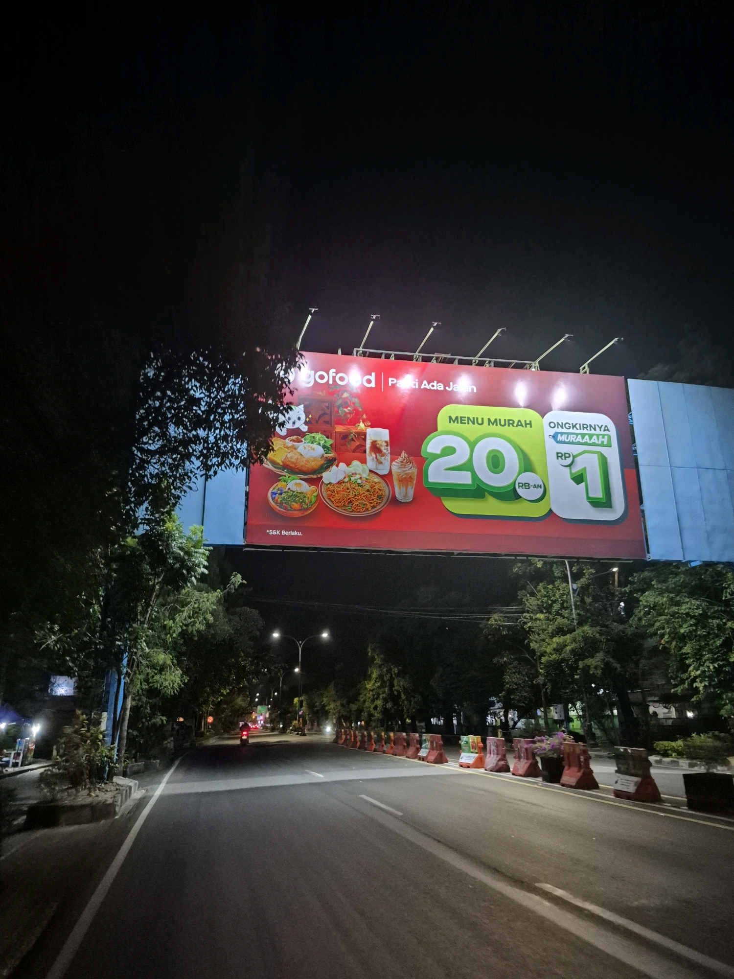 An eye-level medium shot captures a nighttime scene on a paved road in Surakarta, Indonesia, with a large billboard illuminated above. The billboard displays a red background with "gofood" logo in white and promotional text in Indonesian. Images of various Indonesian dishes, including nasi goreng and ayam geprek, are featured. The billboard also highlights a promotion for "MENU MURAH" (cheap menu) for "20" and "ONGKIRNYA MURAAH" (cheap delivery fee) for "RP 1". The road is empty except for a lone motorcycle rider in the distance, illuminated by its headlight, heading away from the viewer. The road is flanked by trees and streetlights. Traffic barriers, some orange and some red, line the right side of the road. The overall atmosphere is dark, with the billboard being the primary source of light.