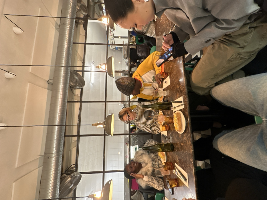 A group of four people are seated at a table. One person in a gray jacket is using a phone. Another wears a yellow jacket. The third person has on a striped shirt, and the fourth is wearing a fur coat. The table has wine bottles, glasses, cutlery, and a plate of food. Overhead, lights hang from the ceiling, near an air duct. The background includes a wall with dark-framed windows.