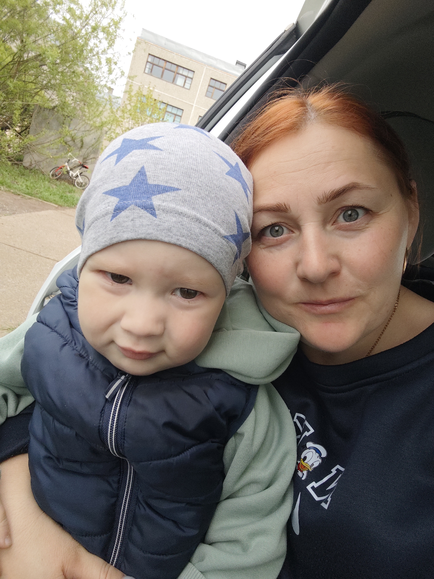 A selfie photograph depicts a woman and a child. The woman has red hair, fair skin, and light-colored eyes. She is wearing a dark-colored sweatshirt with a cartoon character graphic on it. The child is wearing a gray hat with blue stars and a navy blue vest over a light green hooded sweatshirt. The child appears to be held by the woman. The background suggests the photograph was taken inside a vehicle. Outside, there are trees, a building with multiple windows, and a bicycle. The image was taken in Markovskii, Russia.
