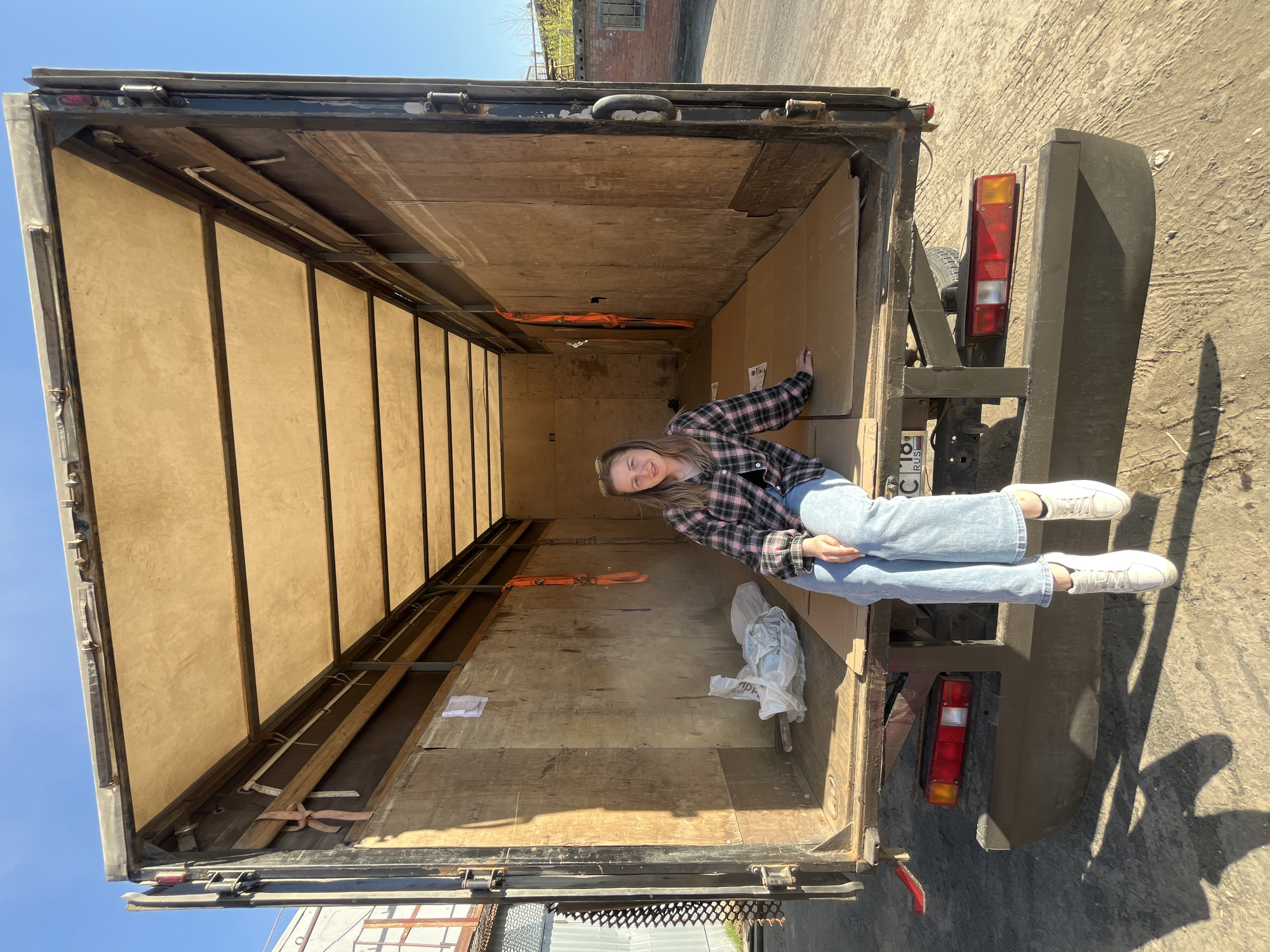 A young woman is sitting in the back of an empty truck. She wears a plaid shirt, light blue jeans, and white sneakers. The interior of the truck is wooden with metal framing. Orange straps are visible inside. A plastic bag is to the left of the woman. The license plate of the truck is visible. The location is Izhevsk, Russia.