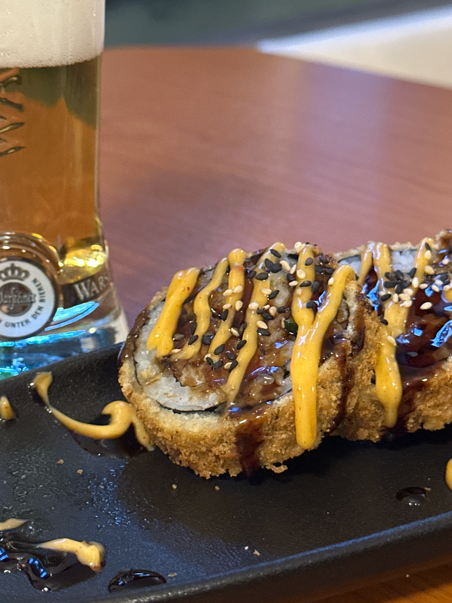 An overhead close-up shot shows a glass of beer and sushi on a black plate. The beer is light brown and foamy, with a label on the glass that reads "WARSTEINER BIEREN UNTER DEN..." The sushi rolls are fried and topped with a creamy orange sauce, dark brown sauce, and black and white sesame seeds. The plate is dark and slightly textured, with some sauce dripped onto it. The background is a wooden table. The image appears to be taken in a restaurant.