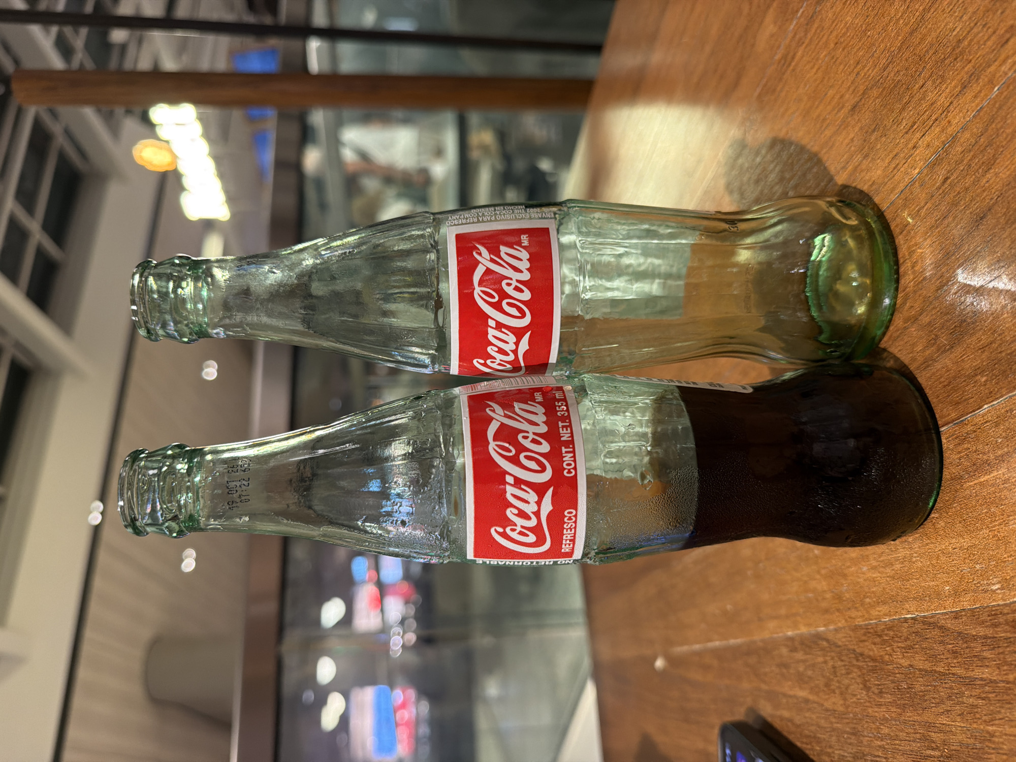 Two glass bottles of Coca-Cola are displayed on a wooden table. The bottle on the left is partially filled with a dark liquid, while the bottle on the right is empty. Both bottles have the iconic red Coca-Cola label. In the background, out-of-focus elements suggest an indoor setting with bright lighting and reflections on glass surfaces. The overall scene conveys a casual and potentially commercial environment. The location is Aventura, United States of America.