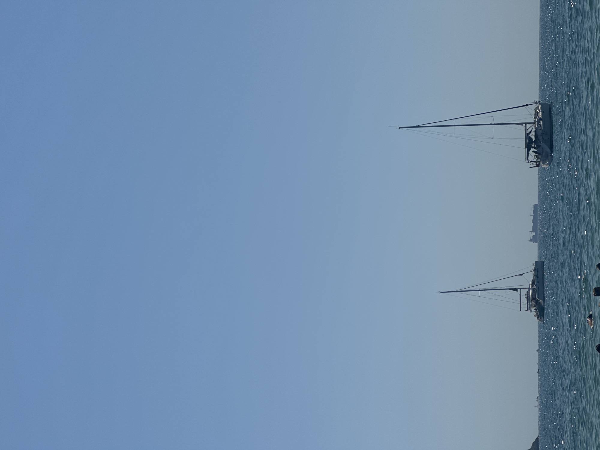 The image depicts a clear, bright day at sea, likely in the Gulf of La Spezia near Lerici, Italy, given the presence of sailboats and distant cargo ships. The sky is a serene, cloudless blue, dominating the upper half of the frame. The sea is a deep blue, with small, shimmering waves catching the sunlight.
In the foreground, partially submerged in the water, are the heads of several individuals, suggesting they are swimming or wading. Their presence indicates a recreational activity taking place.
Further out at sea, two prominent sailboats are visible. The sailboat on the right is closer and larger, with its mast and rigging clearly defined against the sky. It appears to be a monohull sailboat. The sailboat on the left is a catamaran, distinguished by its twin hulls and a wider deck. Both sailboats are anchored or drifting calmly.
In the distance, beyond the sailboats, several large cargo ships are visible on the horizon, suggesting this is a busy shipping lane. A landmass, possibly a coastal hill or island, is dimly visible on the far left of the image. The overall scene conveys a sense of peacefulness and leisure on a summer day.
