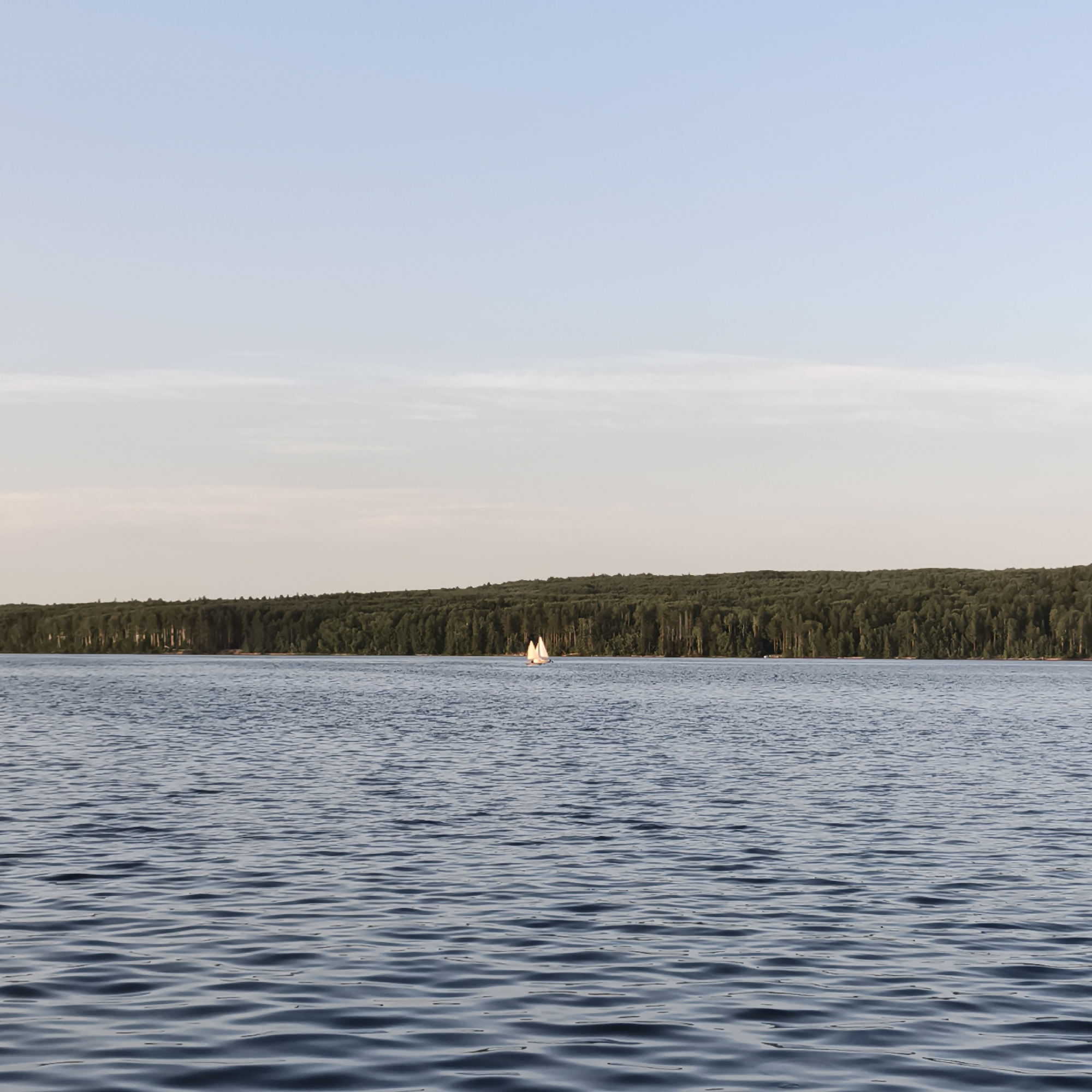 A sailboat with two sails is visible in the middle distance of a wide, blue lake. The lake's surface is rippled, suggesting a gentle breeze. In the background, a dense, dark green forest stretches along the horizon, with the tops of the trees creating an irregular line against the pale sky. The sky itself is a soft blue, with faint, wispy clouds near the horizon. The overall impression is a serene natural landscape. This scene is likely located on the Kama River reservoir near the city of Chaikovsky, Russia.