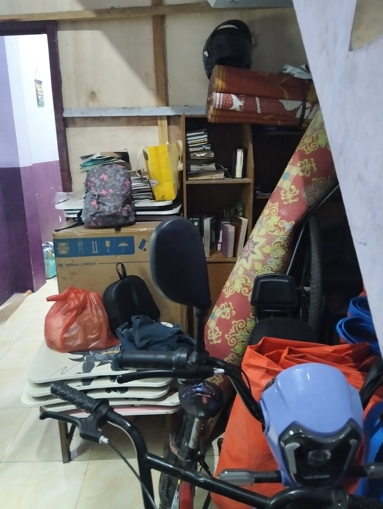 The image depicts a cluttered indoor storage area, likely a room or garage, in the city of Medan, Indonesia. The scene is filled with various objects, suggesting a disorganized environment.
Prominently displayed in the foreground is the front section of a bicycle, including its handlebars and a light blue front fairing. To the left of the bicycle's handlebars, on a stack of light-colored tables, is a bright orange plastic bag. To the right of this bag is a black backpack, and further right, a pile of dark blue fabric. Behind these items, a large cardboard box with shipping symbols is visible.
In the background, a shelving unit is packed with books, papers, and other assorted items. On top of the shelving unit, several rolled-up mats are stacked, topped by a black helmet. A large rug with a red and yellow patterned design is propped up against the wall to the right, partially obscuring a bicycle wheel. The walls appear to be made of wood paneling and a light-colored material.
The overall impression is one of a space used for storage, with items piled and stacked in a somewhat haphazard manner. The lighting suggests an indoor environment, possibly with natural light filtering in from an unseen source.
