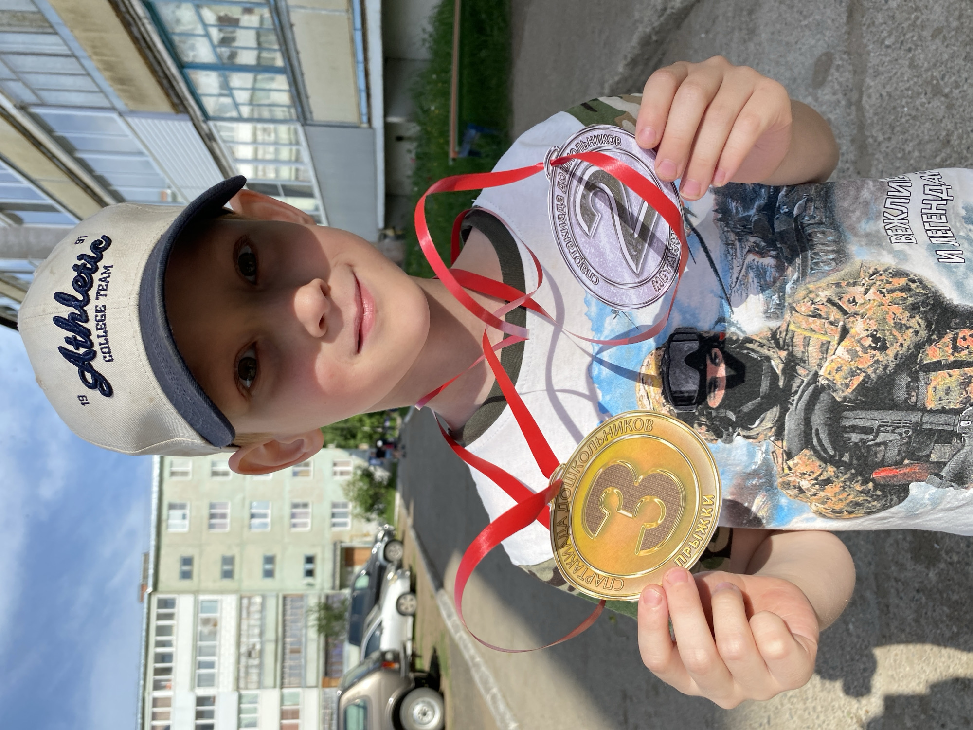 A young boy stands outdoors, holding two medals with red ribbons. One medal is silver with the number "2", and the other is gold with the number "3". He is wearing a baseball cap that says "Athletic COLLEGE TEAM" and a t-shirt featuring military figures. The backdrop includes an apartment building and parked cars, suggesting an urban area. The location is Noviy, Russia, as inferred from the Russian text on the medals.