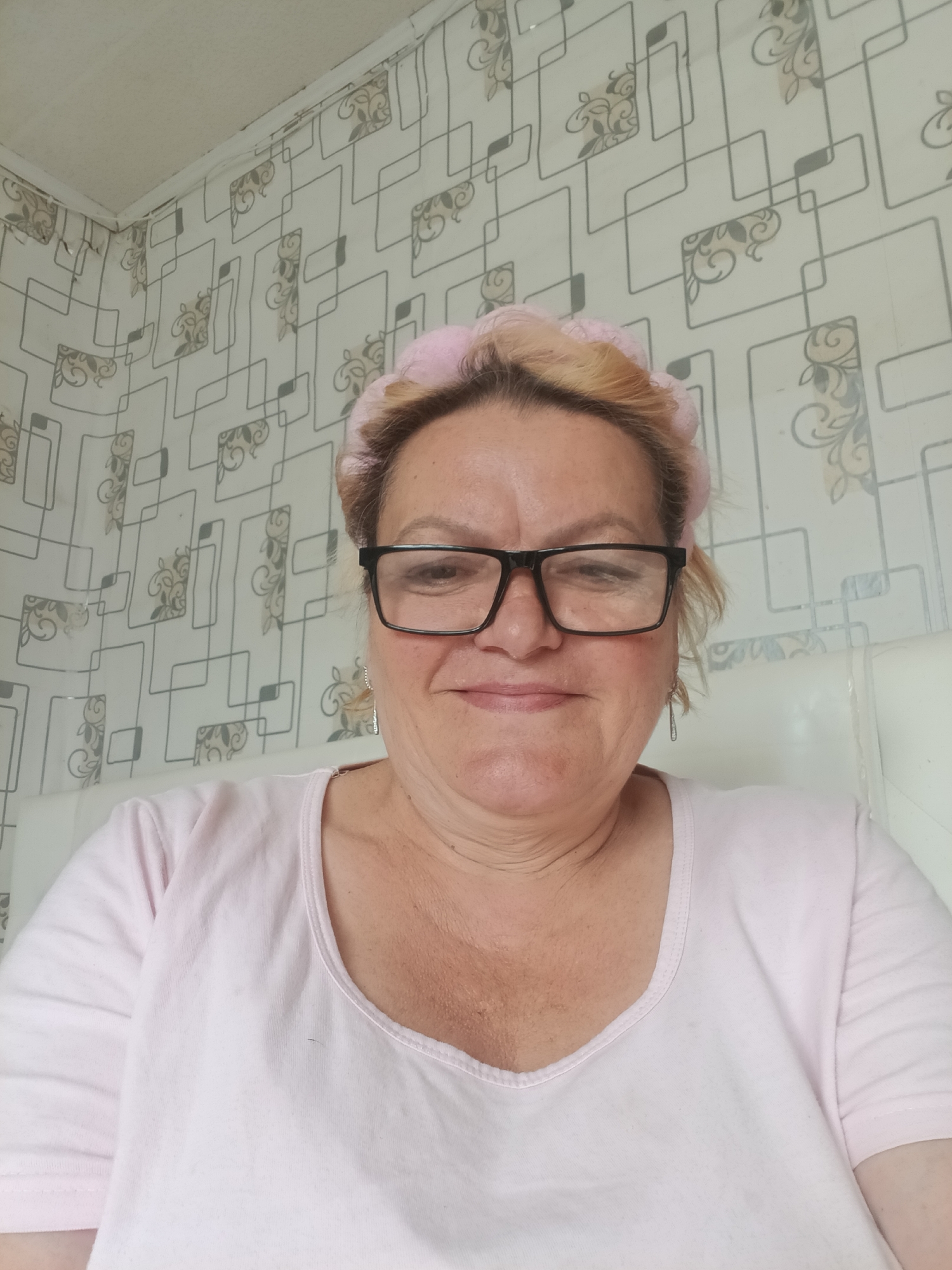 A close-up selfie shows a middle-aged woman looking directly at the camera with a gentle smile. She has light-colored hair, which is held back by a pink, possibly fluffy, headband or headscarf. She wears black-framed rectangular glasses and a light pink, short-sleeved t-shirt with a scoop neck. Small, metallic dangle earrings are visible.
The woman is positioned indoors, likely in a room in Красный Маяк, Russia. The background features light-colored wallpaper adorned with a repeating pattern of interlocking dark grey geometric lines and interspersed lighter brown or grey floral/scroll motifs. In the upper left quadrant, the corner where the wall meets the ceiling is visible, featuring a white trim piece. A plain white or light-colored surface, possibly a headboard or furniture, is visible behind her right shoulder. The lighting appears soft and even, suggesting a daytime setting. No discernible text is visible in the image.