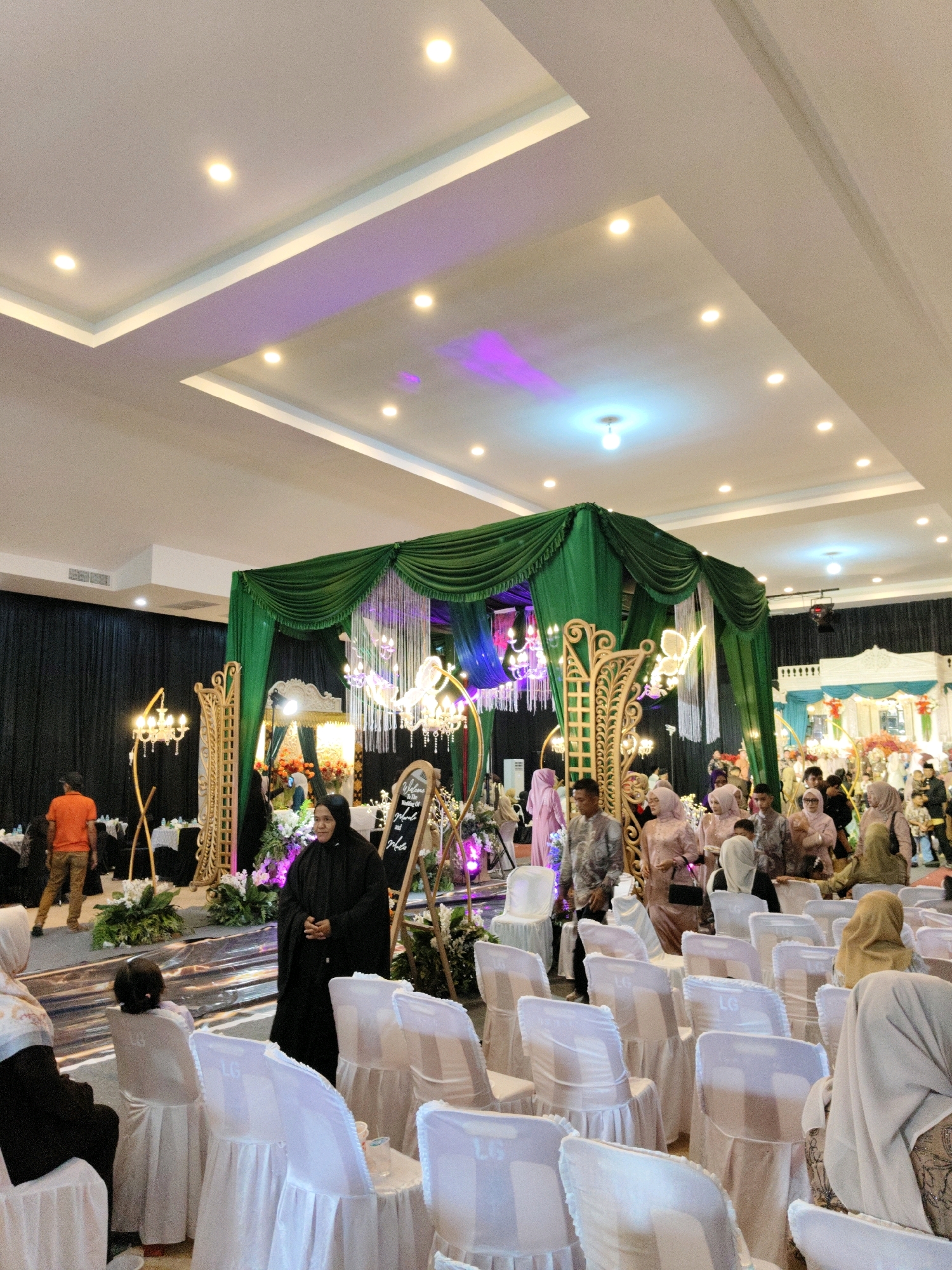 The image depicts an indoor event, possibly a wedding reception or a similar formal gathering, set up in a banquet hall. The hall features a high, white ceiling with recessed lighting and some decorative purple and blue colored lights. A prominent stage or decorated area is visible in the background, draped with green fabric and adorned with crystal chandeliers and floral arrangements. A dark curtain forms the backdrop for this stage.
In the foreground and mid-ground, rows of white-covered chairs are arranged, some with small tables. Several people are present, dressed in formal attire, including women wearing hijabs and traditional Indonesian clothing in various colors like pink, beige, and black. One woman in the mid-ground, wearing a black hijab and dress, is facing the camera and appears to be addressing the viewer. Another woman in a black outfit is seated in the lower left, partially visible. A man in an orange shirt and khaki pants is seen on the left side, walking away from the camera. The overall atmosphere suggests a celebratory event. The context provided indicates this scene is located in Lhokseumawe, Indonesia.