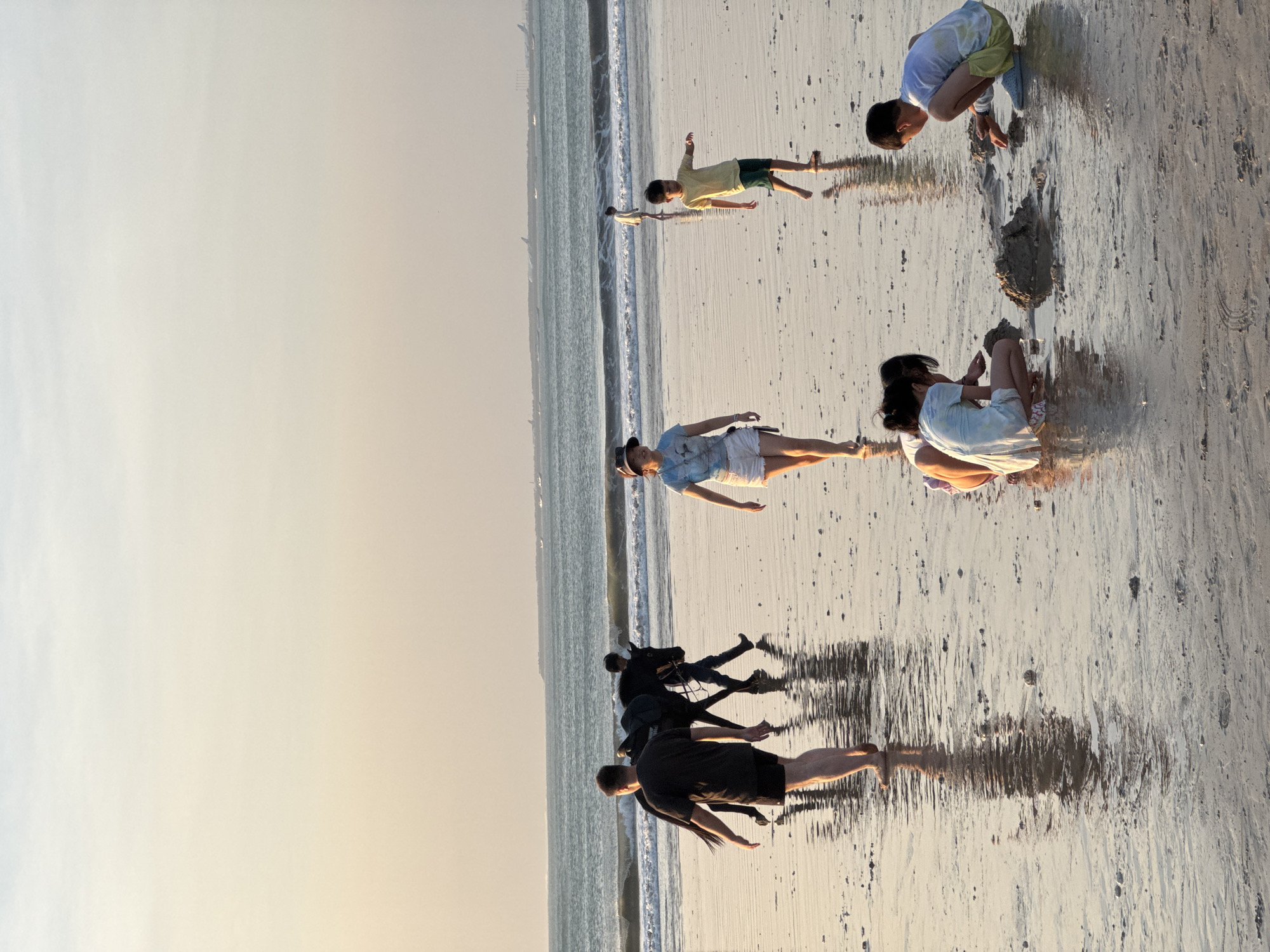 This is a photograph taken at the beach in Kuta Selatan, Indonesia, during what appears to be sunset. The scene depicts several individuals engaged in various activities on the wet sand near the ocean.
In the foreground, two children are crouched down, actively playing with sand and building something, possibly a sandcastle. One child is wearing a light blue tie-dye t-shirt and yellow shorts, while the other is wearing a light blue and white tie-dye dress.
Further back, a woman with a hat and a light blue t-shirt with white shorts is walking across the beach. To her right, a young boy in a yellow t-shirt and green shorts is also walking.
In the middle ground, two people are walking, one of them leading a dark-colored horse. The horse is saddled and appears to be walking calmly. The individuals are dressed in casual beachwear.
The background shows the ocean with gentle waves rolling onto the shore. The sky is a soft gradient of light blue and peach, indicative of sunset. In the far distance, faint shapes of what appear to be buildings or structures are visible on the horizon, along with some boats or ships. The wet sand reflects the light and the figures present, creating a shimmering effect. The overall atmosphere of the image is tranquil and suggests a leisure activity at the beach.