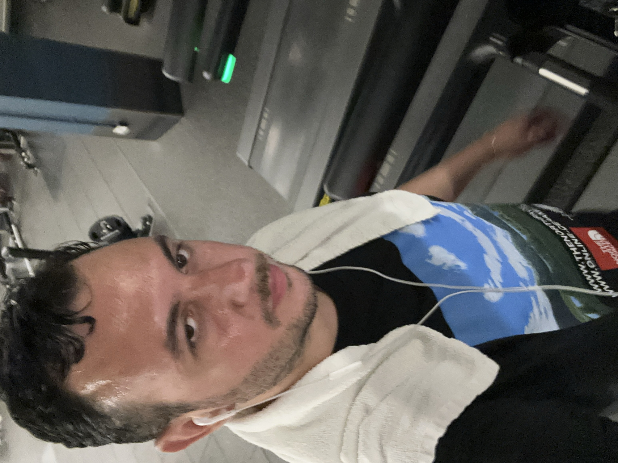 A selfie shows a sweaty, light-skinned man with dark, curly hair and a trimmed beard, wearing a black T-shirt partially covered by a white towel, presumably after a workout. Earphones are plugged in. The background includes a treadmill and other gym equipment. Based on the T-shirt logo, the location may be a gym somewhere in London, United Kingdom.
