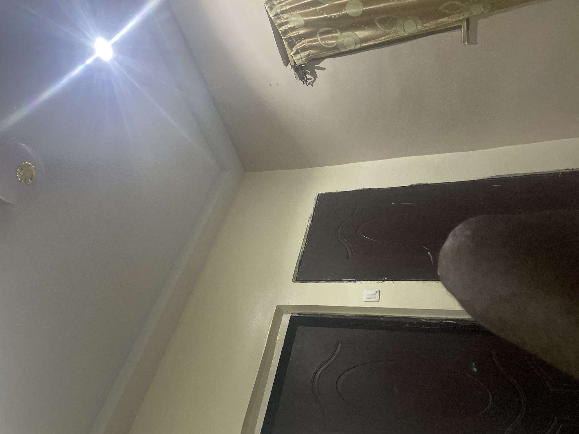 The image depicts an interior scene, possibly a room in Jalingo, Nigeria. A bright light source is prominent in the upper right quadrant of the image, creating lens flare and illuminating the ceiling. The ceiling is white with a decorative molding. On the left side of the image, two dark brown doors are visible, close to each other. One door has a white light switch mounted on the wall next to it. A patterned curtain, gold in color, is partially visible on the right side of the image, hanging from a rod. In the foreground, a blurred, dark, curved object, possibly a leg or part of a person, occupies the lower central portion of the frame. The walls are painted in a light, neutral color. The overall lighting suggests it might be nighttime or a dimly lit interior with a strong artificial light source.
