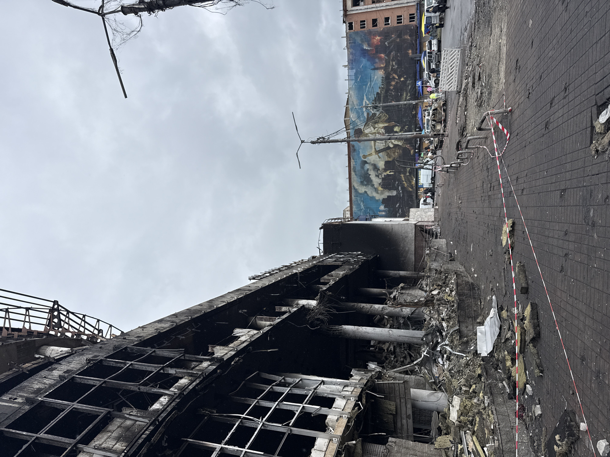 The image shows the aftermath of an apparent bombing in Kyiv, Ukraine. A building is heavily damaged, with its facade charred and significant structural damage visible. Debris litters the ground. A large billboard depicting a historical scene is visible behind the damaged building. Cars and emergency vehicles are parked along the street, which is paved with dark bricks. Red and white tape is used as a barrier, indicating a restricted zone. The sky is overcast.