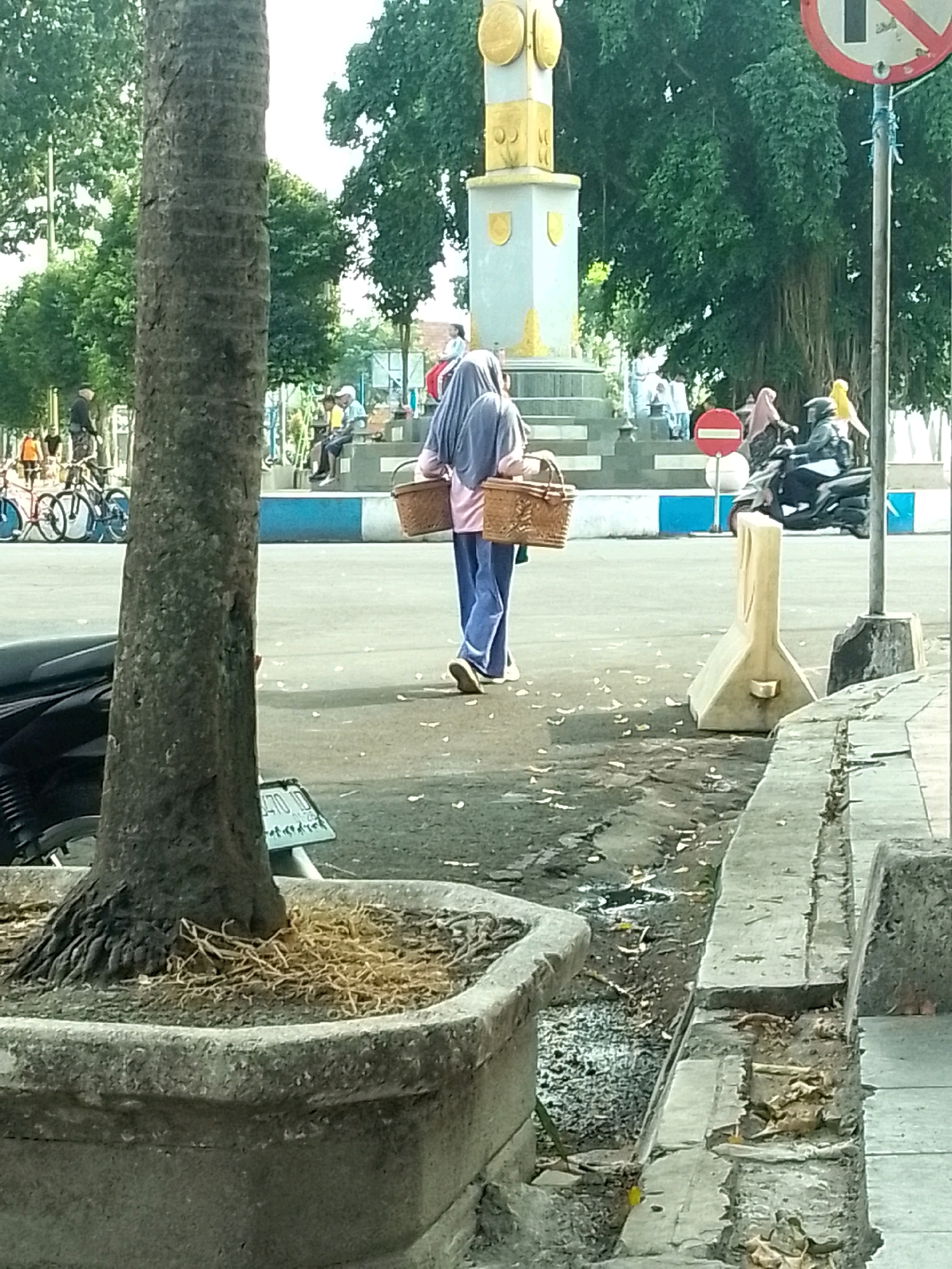 The image captures an outdoor scene in Jember, Indonesia, likely a public space such as a park or plaza. A person wearing a hijab and blue trousers is walking away from the camera, carrying two woven baskets, one in each hand. In the background, a monument with a gold-colored top stands tall amidst lush green trees. Several other individuals are visible in the background, some sitting and others walking. There are bicycles parked to the left of the frame, and a motorcycle is present in the mid-ground. A concrete barrier and a drain are prominent in the foreground, suggesting an urban or semi-urban environment. The overall impression is a slice of daily life in a moderately busy public area.