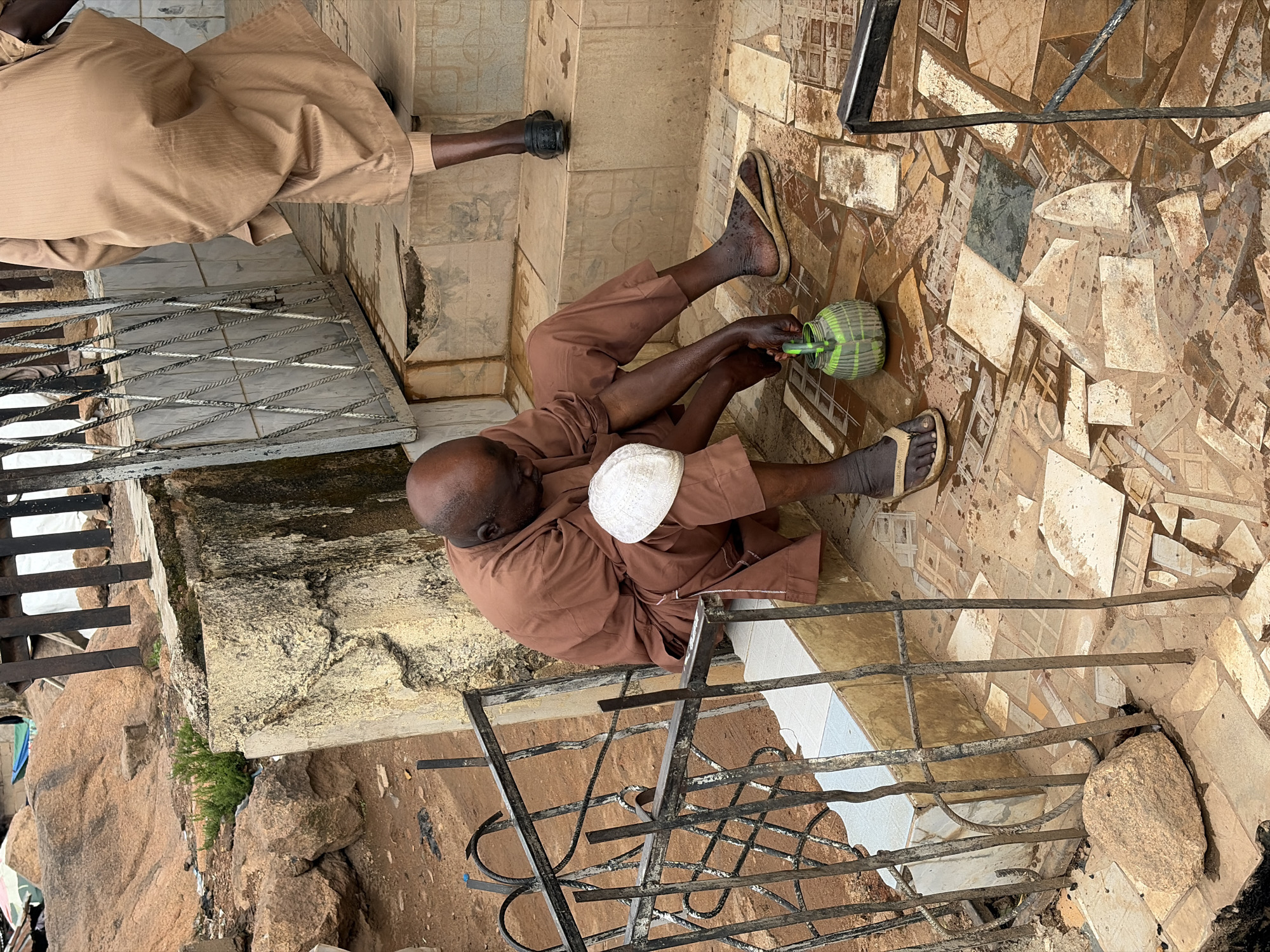 The image shows two men in a semi-outdoor setting, likely in Jos, Nigeria. One man, wearing a tan robe and a white cap, is seated on the ground with his legs extended. He is holding a green and white watering can with his right hand and appears to be tending to something. His left hand is resting on his lap. The other man, wearing a similar tan robe, is seated higher up on a step, with his legs bent. His legs are visible from the knees down, and he is wearing sandals.
The ground is paved with irregularly shaped tiles in shades of brown and beige, some of which are cracked or missing. A metal railing runs along the foreground, obscuring parts of the scene below. In the background, there are rough-hewn rocks and vegetation, suggesting a somewhat unkempt or natural environment. The lighting suggests it is daytime. The overall impression is of a moment of rest or routine activity. There is no visible text in the image.