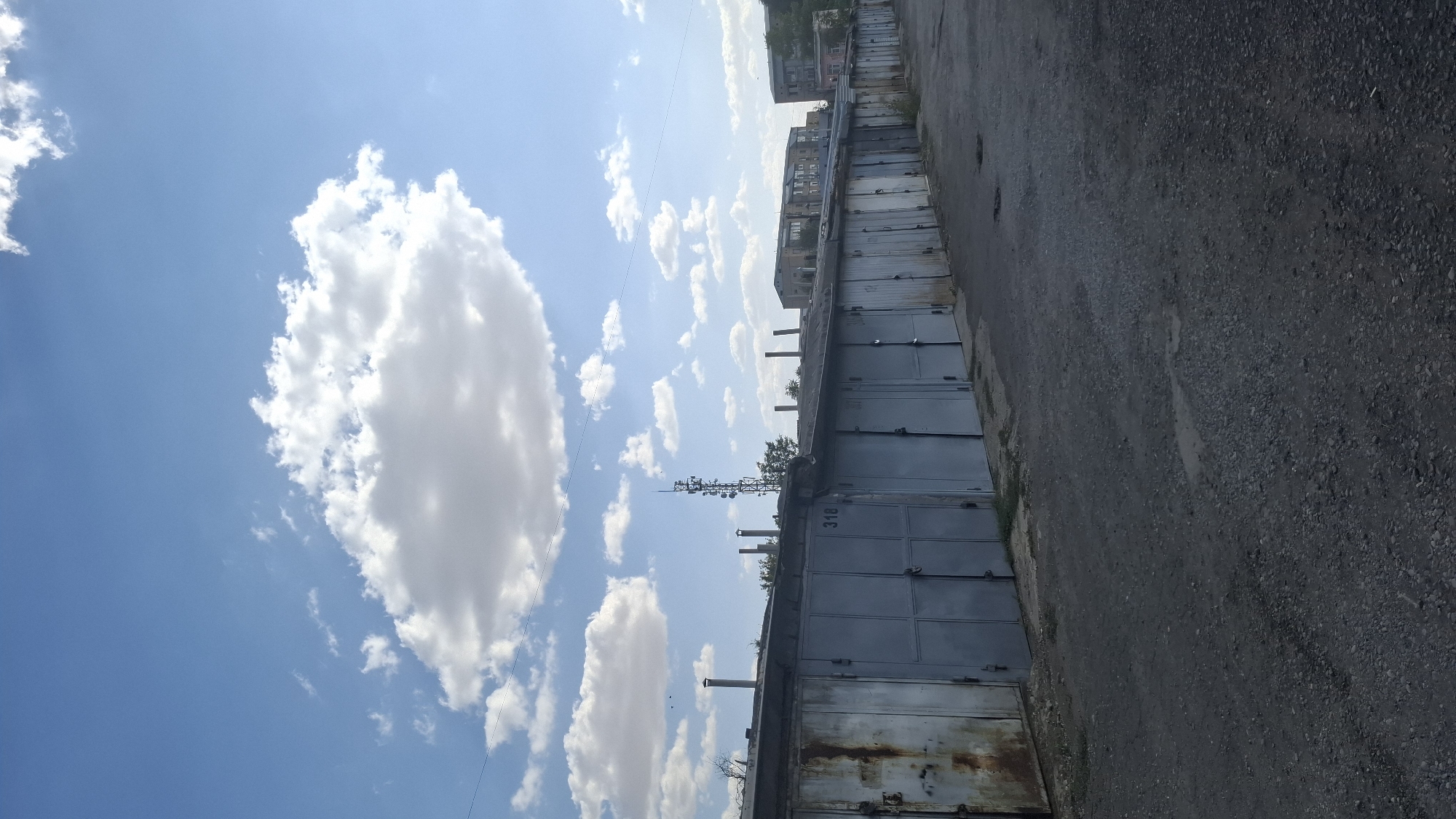 A daytime, vertical, eye-level shot captures a row of identical metal garages situated on a gravel surface. The garages are painted a dull gray, with some showing signs of rust and wear. Garage number 318 is clearly visible on one of the doors. The sky above is a bright blue, dotted with fluffy white clouds, indicating fair weather. In the background, a tall telecommunications tower stands prominently, and several residential buildings are visible in the distance. The scene is likely located in Shymkent, Kazakhstan, given the architectural style of the garages and buildings, common in post-Soviet urban environments. The overall impression is one of a utilitarian, working-class neighborhood.