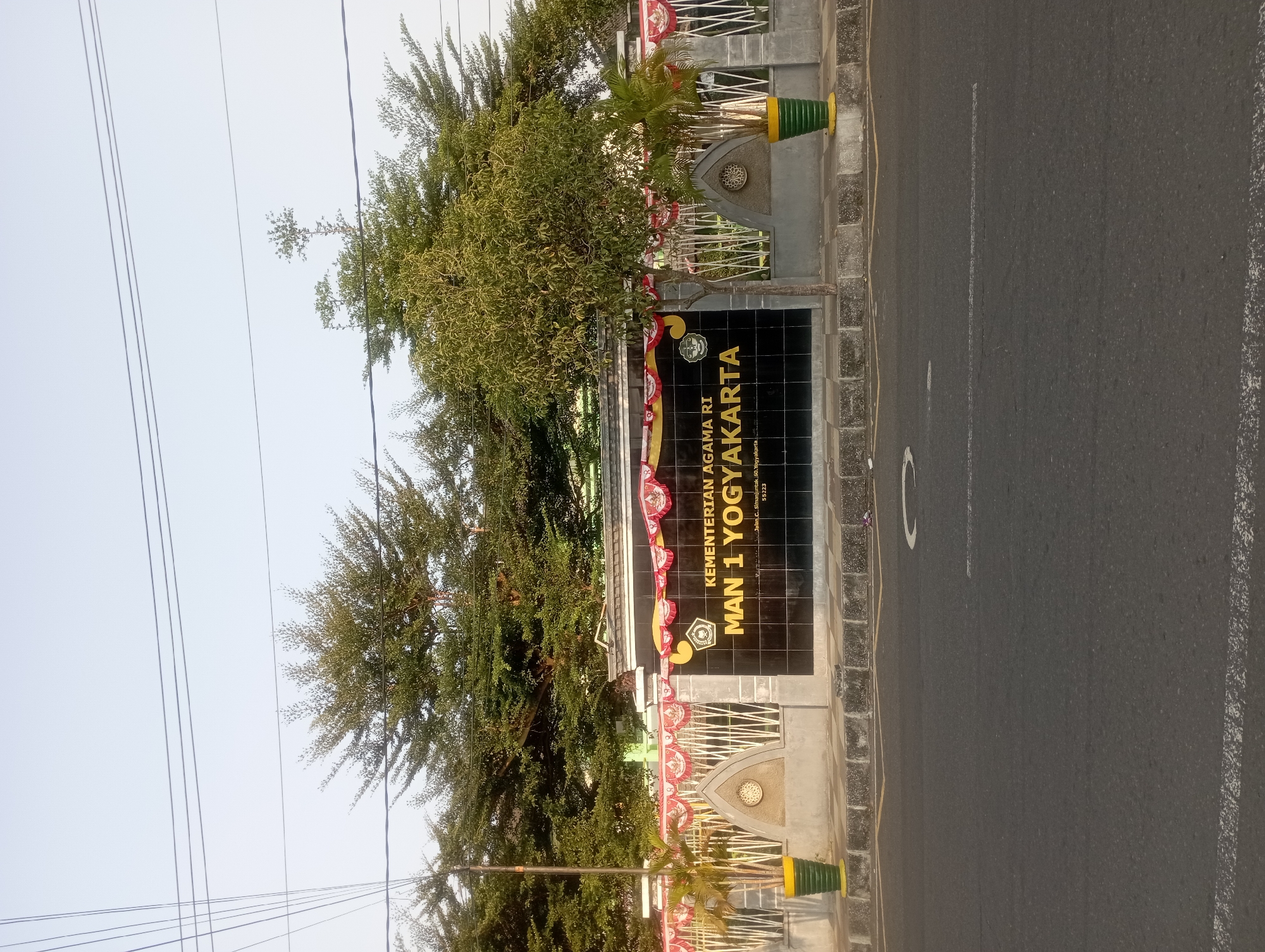 The image shows the entrance to MAN 1 Yogyakarta, a public Islamic senior high school located in Yogyakarta, Indonesia. The school's name is prominently displayed on a black tiled sign with yellow lettering. The sign reads "KEMENTERIAN AGAMA RI MAN 1 YOGYAKARTA" and includes an address and postal code. The entrance is flanked by a fence and decorated with red and white banners. Two potted plants are visible, one on each side of the entrance. Behind the entrance, a row of trees can be seen. The scene is captured from a street-level perspective, with a road in the foreground. The sky is clear and the lighting suggests it is daytime, likely morning or late afternoon due to the low angle of the sun. There are no people visible in the image.
