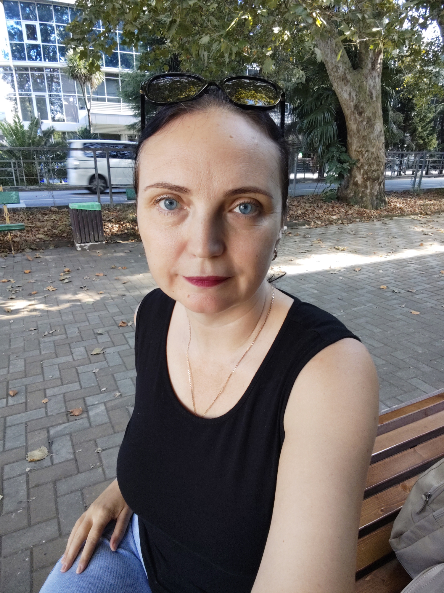 A woman with blue eyes and dark hair, wearing a black tank top and sunglasses perched on her head, poses for a selfie outdoors. She is seated on a wooden bench in a paved area with scattered autumn leaves. Behind her, a building with reflective windows and lush green trees are visible. A white van is seen driving on a road in the background, suggesting an urban or suburban setting. The overall atmosphere is casual and relaxed, possibly in a park or public space within the city of Sochi, Russia.