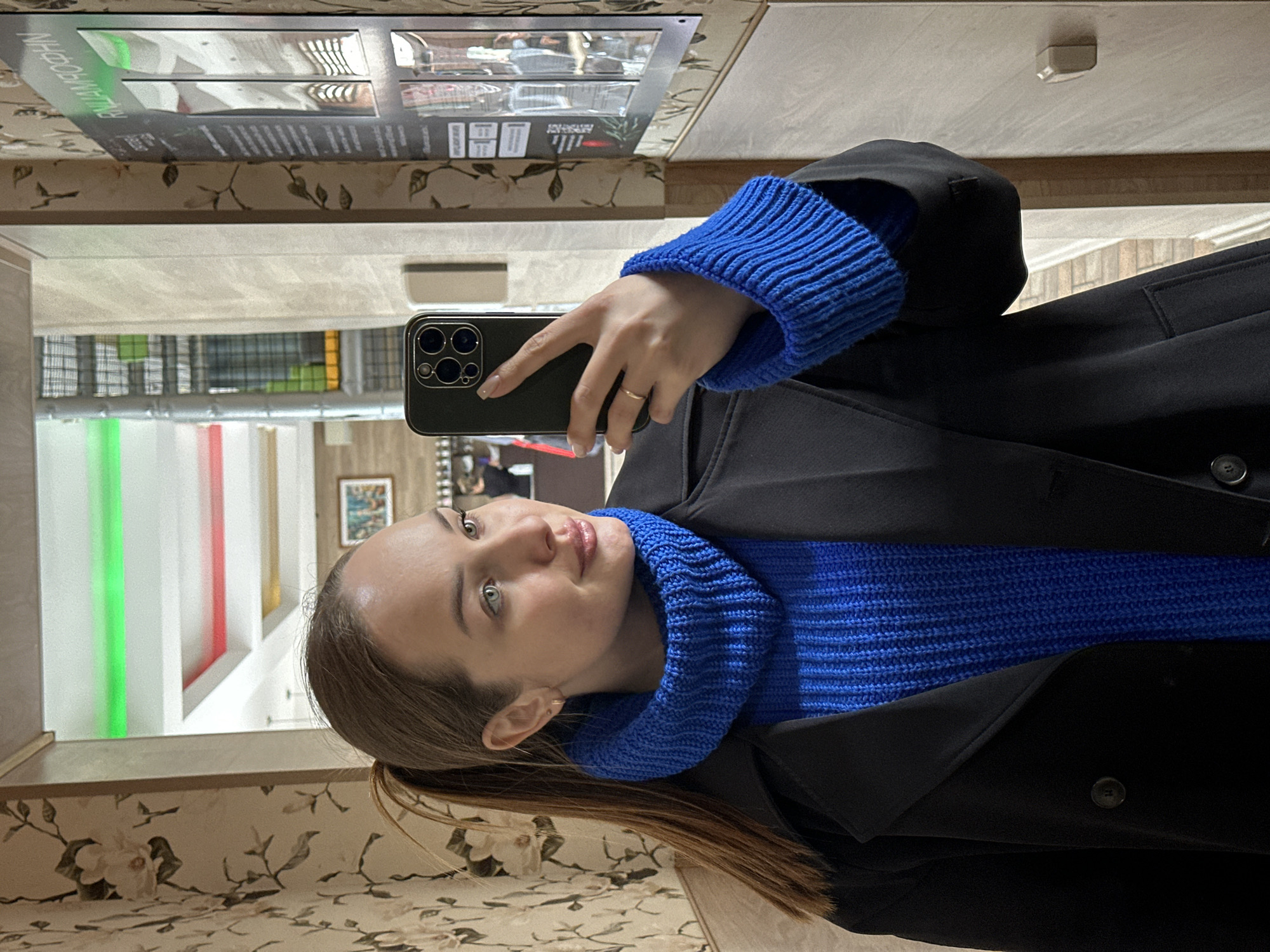 A woman is taking a selfie in a mirrored hallway. She is wearing a blue turtleneck sweater and a black coat. She is holding a black smartphone. The background shows a hallway with green and red vertical stripes, a painting on the wall, and wallpaper with a floral pattern. Reflected on the top of the mirror is a poster displaying the word “АЛМАЗООХИН” in green. The location is Chaikovsky, Russia, based on the text on the poster.